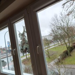 Usługi Maria Iwaszko - Trzyczęściowe, białe okno PCV z widokiem na zewnątrz: drzewa, trawnik i budynki w pochmurny dzień. Widoczne detale okna, w tym klamki i uszczelki.