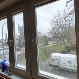 Usługi Maria Iwaszko - Widok z okna po umyciu, z widocznymi smugami na szybach. Na parapecie dekoracje. Za oknem budynki, drzewa i samochód firmy sprzątającej.