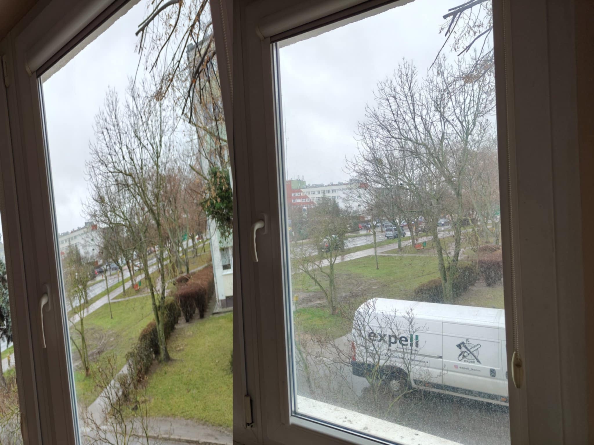 Widok z okna na zewnątrz, po umyciu. Na szybie brak smug, w tle budynki, drzewa i samochód firmy sprzątającej. Czyste okno z białymi ramami.