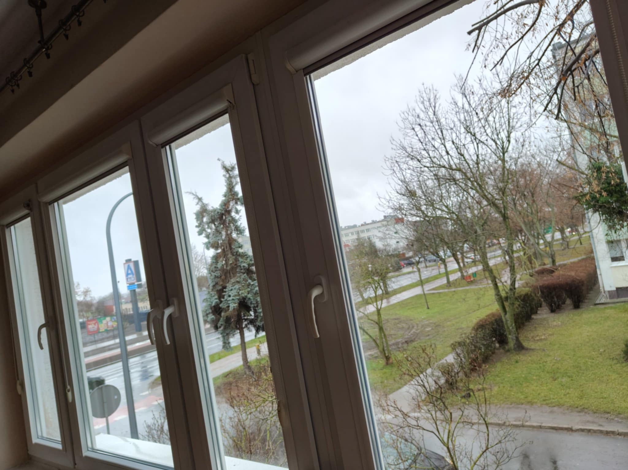 Trzyczęściowe, białe okno PCV z widokiem na zewnątrz: drzewa, trawnik i budynki w pochmurny dzień. Widoczne detale okna, w tym klamki i uszczelki.