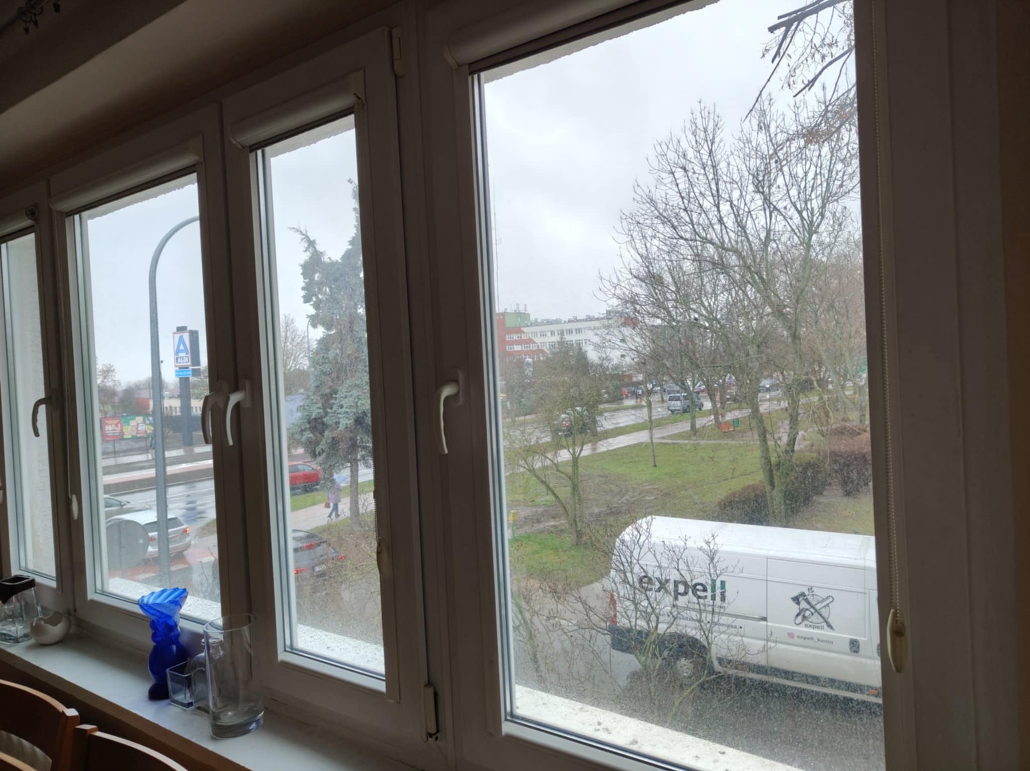 Widok z okna po umyciu, z widocznymi smugami na szybach. Na parapecie dekoracje. Za oknem budynki, drzewa i samochód firmy sprzątającej.