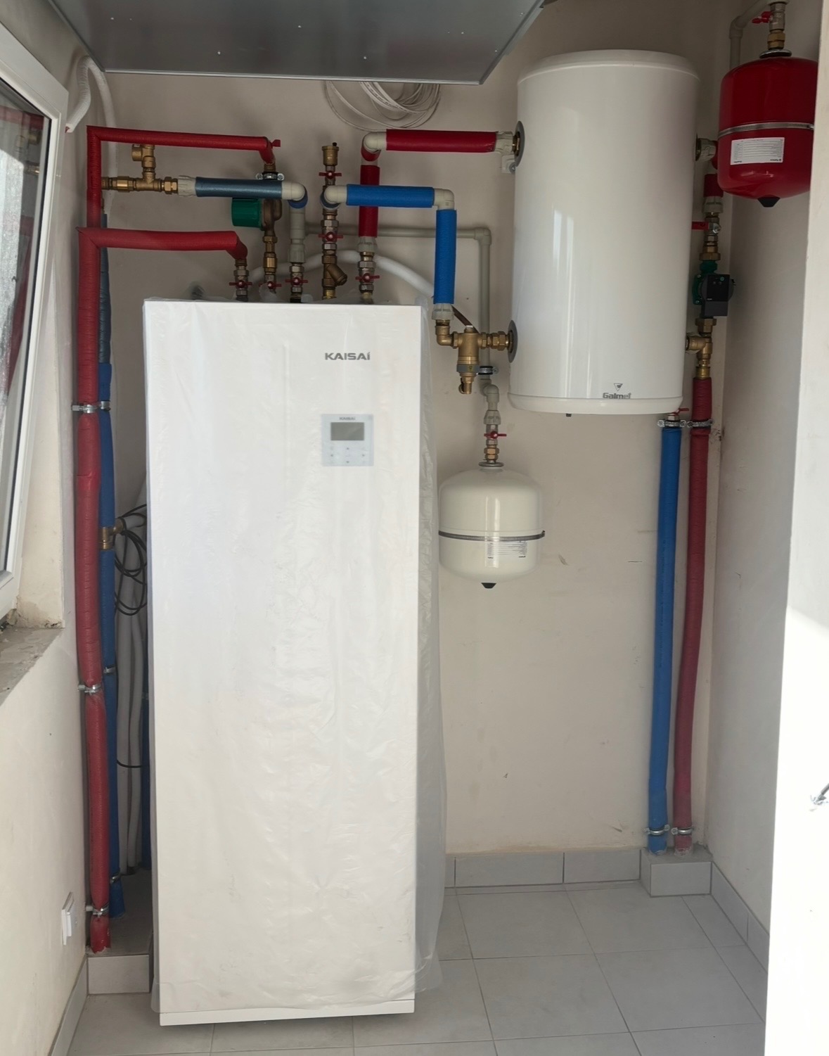 Biała pompa ciepła Kaisai z podłączonymi rurami (czerwone, niebieskie) i zbiornikiem. Widoczne elementy instalacji grzewczej w pomieszczeniu z szarą podłogą.