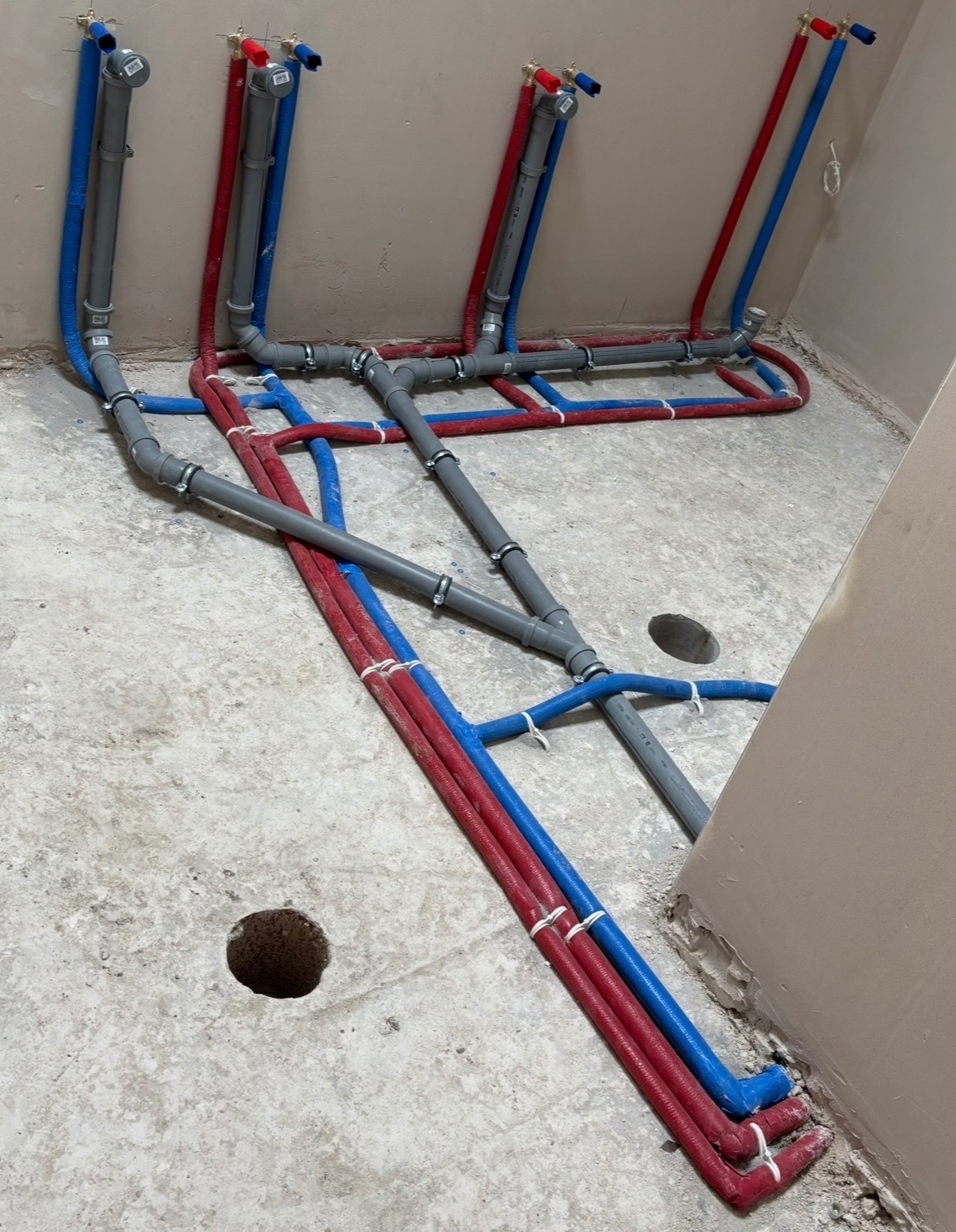 Rozłożona instalacja wodna z rur w kolorach niebieskim i czerwonym, połączonych szarymi elementami. Widoczne otwory w podłodze i fragment ściany.