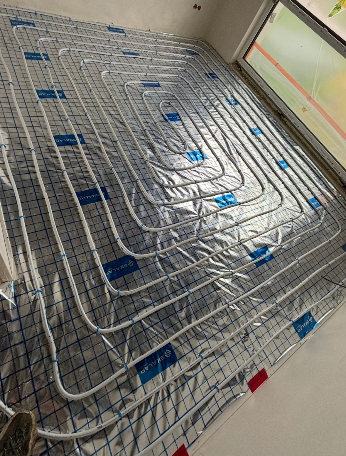 Białe rury ogrzewania podłogowego ułożone na srebrnej folii z niebieską siatką, widoczne z góry pod kątem. Rury układają się w pętle, blisko okna.