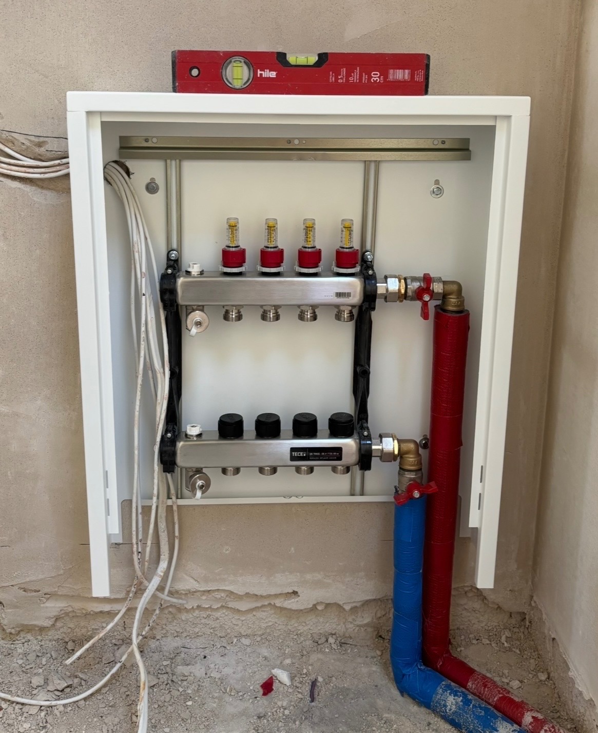 Rozdzielacz ogrzewania podłogowego TECE w białej szafce, z poziomnicą Hile na górze. Czerwone i niebieskie rury prowadzą do instalacji.