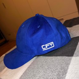 FabriWorks Wiktor Kania - Niebieska czapka z daszkiem z białym haftowanym logo 'CFT' na boku, leżąca na szaro-białej kraciastej tkaninie. Widoczne szwy i faktura materiału.