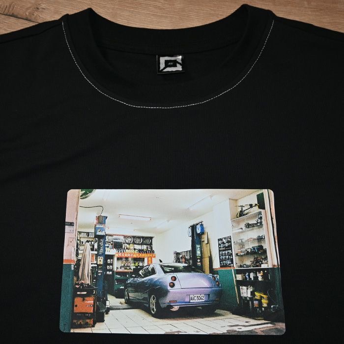 Czarna koszulka z nadrukiem zdjęcia fioletowego samochodu w warsztacie, widoczna metka i białe przeszycia wokół szyi. Ujęcie z bliska, na płaskiej powierzchni.