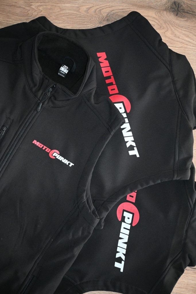 Czarna kamizelka i bluza z nadrukiem 'Moto Punkt' w kolorze czerwono-białym, leżące na drewnianym podłożu. Widoczne detale zamków i metki.