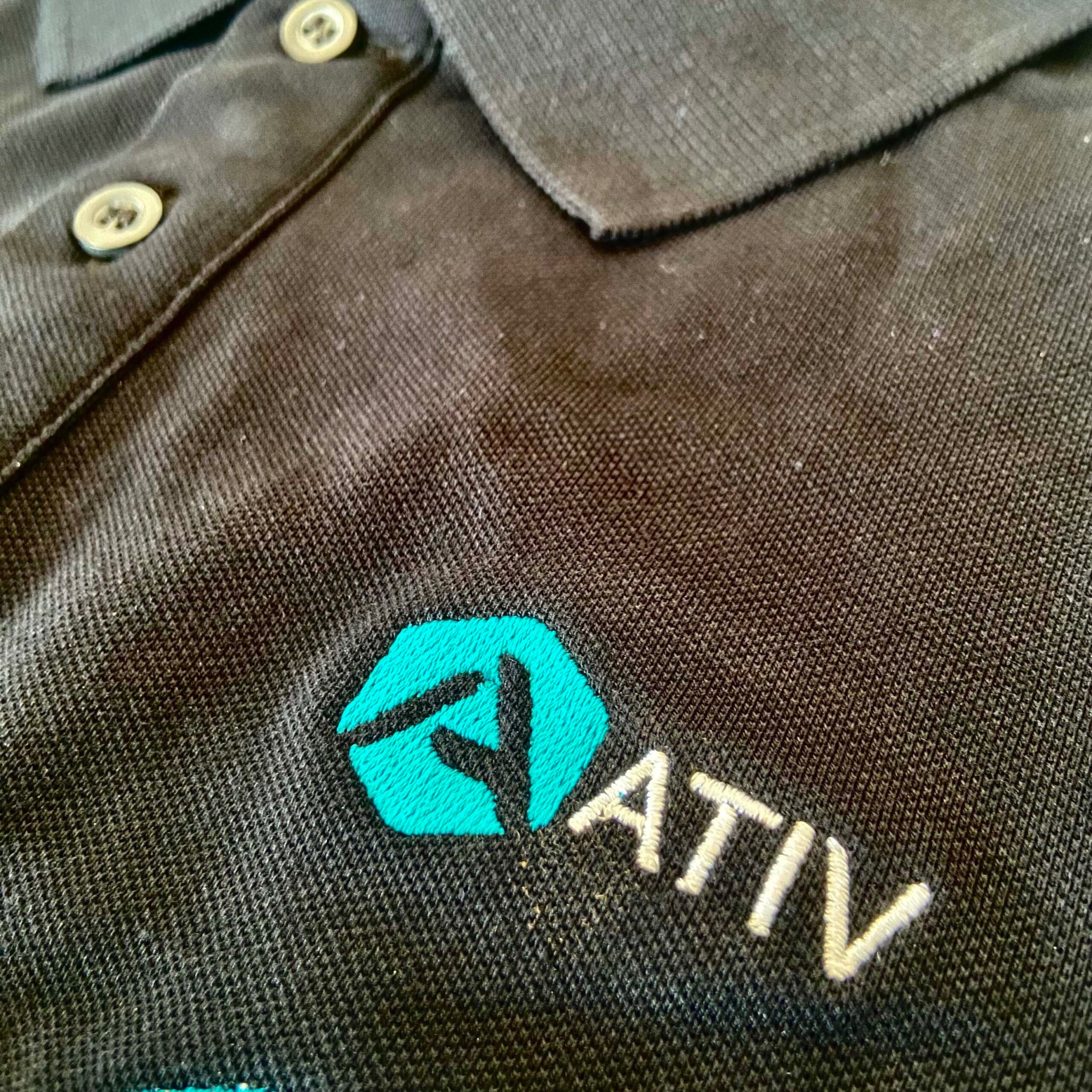 Czarne polo z haftowanym logo 'ATIV' w kolorze turkusowym i białym, widoczne guziki i faktura materiału. Detal haftu firmowego na odzieży.