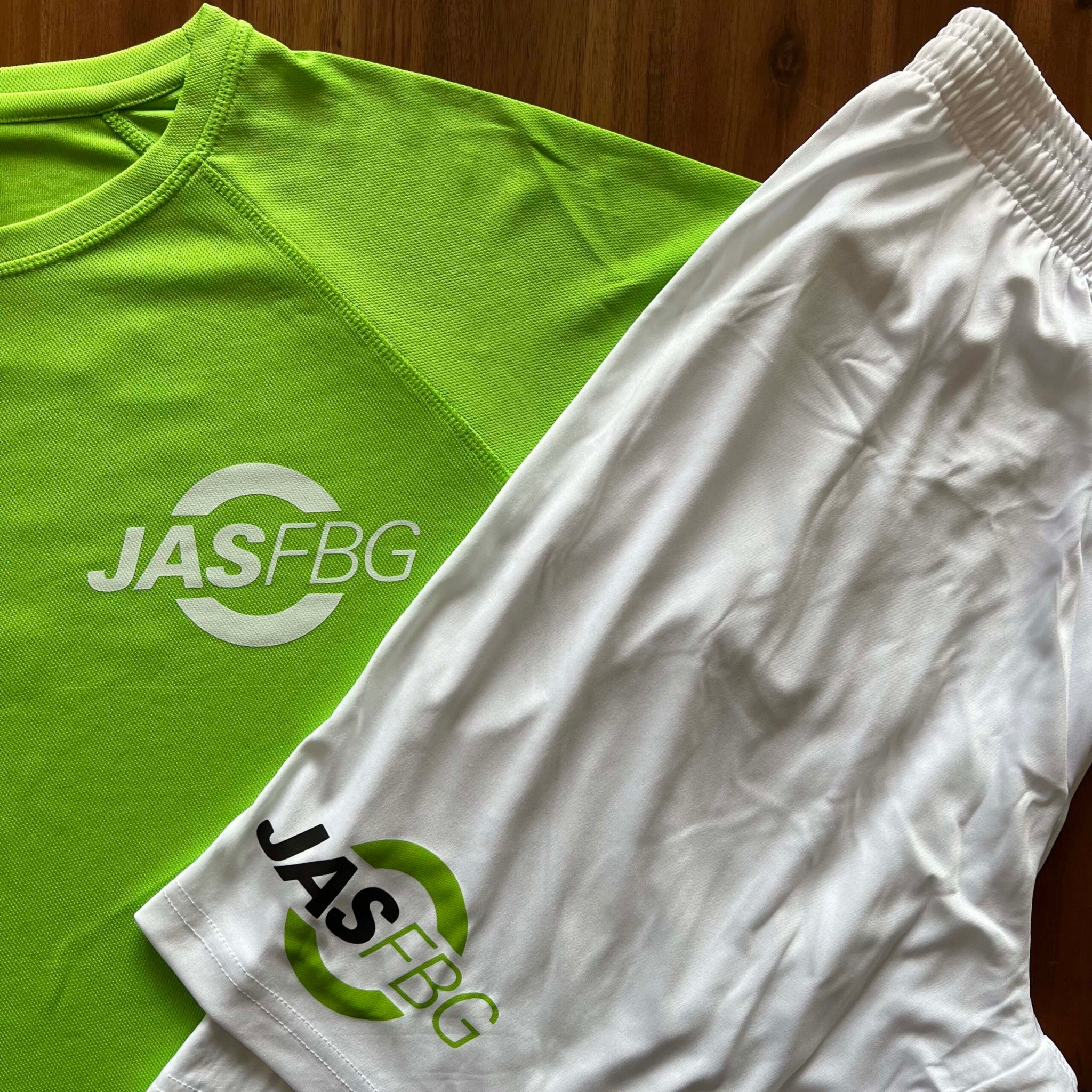 Jasnozielona koszulka sportowa z białym nadrukiem 'JASFBG' i białe spodenki z zielono-czarnym nadrukiem, leżące na drewnianym tle.