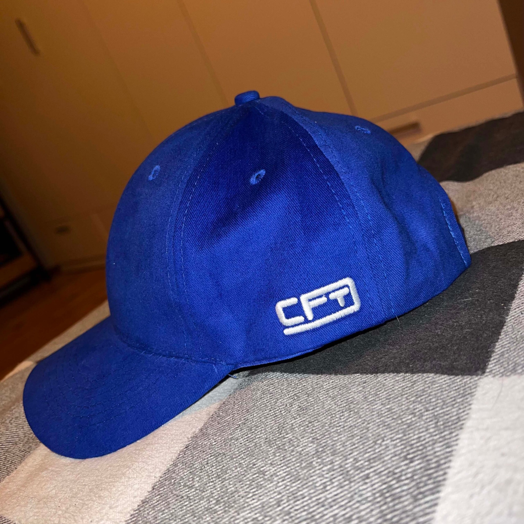 Niebieska czapka z daszkiem z białym haftowanym logo 'CFT' na boku, leżąca na szaro-białej kraciastej tkaninie. Widoczne szwy i faktura materiału.