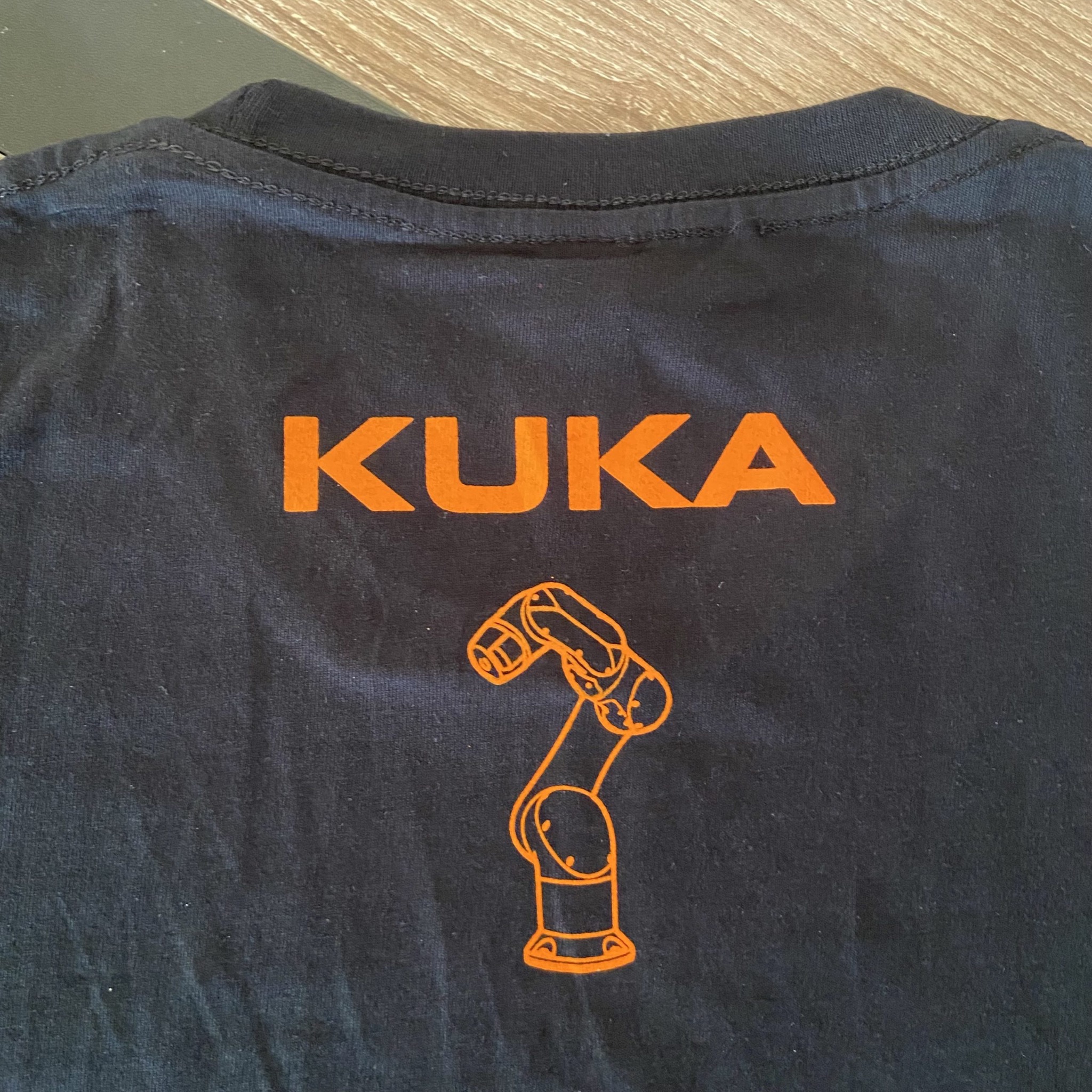 Ciemna koszulka z pomarańczowym nadrukiem: logo KUKA i schematyczny robot przemysłowy. Widok z tyłu, blisko kołnierzyka. Materiał lekko pognieciony.