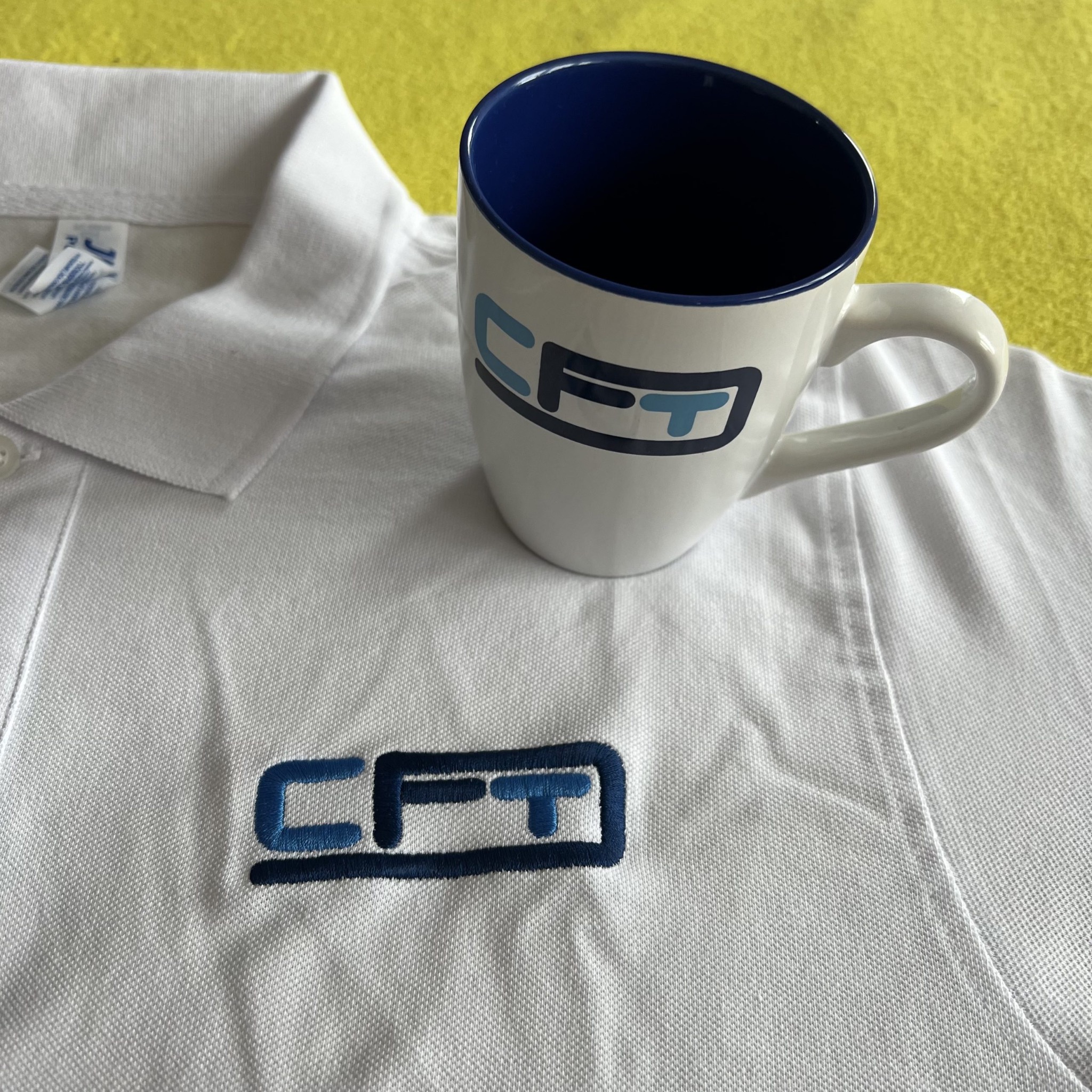 Biała koszulka polo z haftowanym logo 'CFT' w kolorze niebieskim, obok biały kubek z tym samym logo na żółtym tle. Precyzyjne wykonanie haftu.