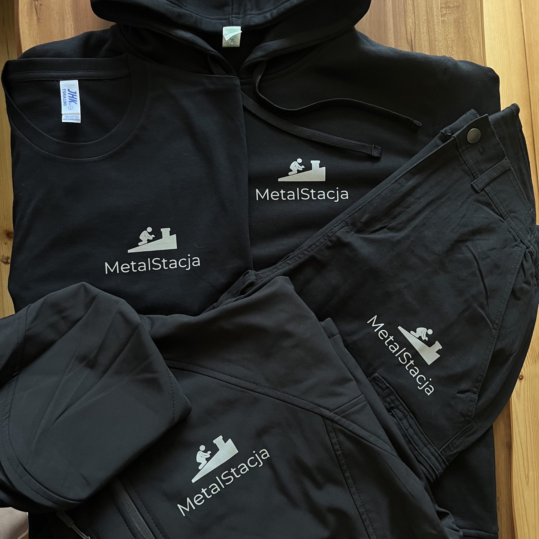 Czarna odzież robocza z logo 'MetalStacja': bluza z kapturem, kurtka, spodnie i koszulka. Ułożone na drewnianym tle. Widoczne metki na koszulce.