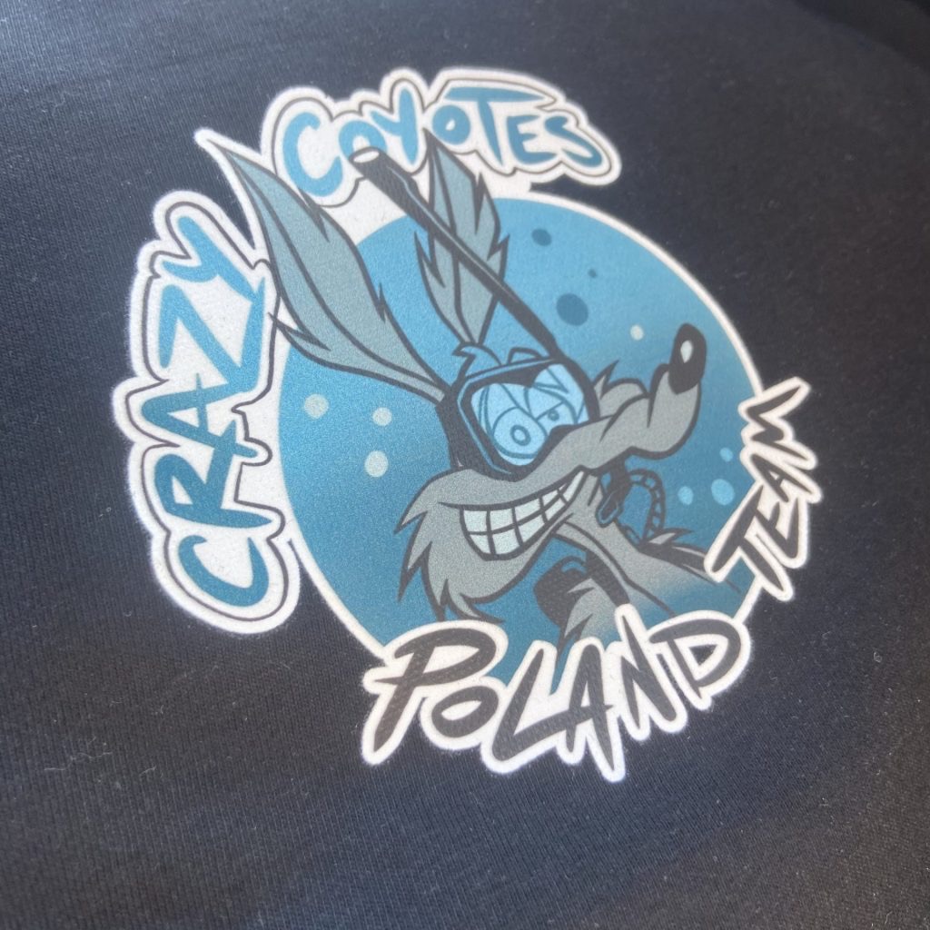 Czarna koszulka z nadrukiem wesołego kojota w okularach do nurkowania na tle błękitnego koła. Napis 'Crazy Coyotes Team Poland'.