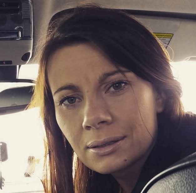 Portret kobiety z brązowymi włosami, siedzącej w samochodzie, z delikatnym uśmiechem i naturalnym makijażem, uchwycony w jasnym, dziennym świetle.