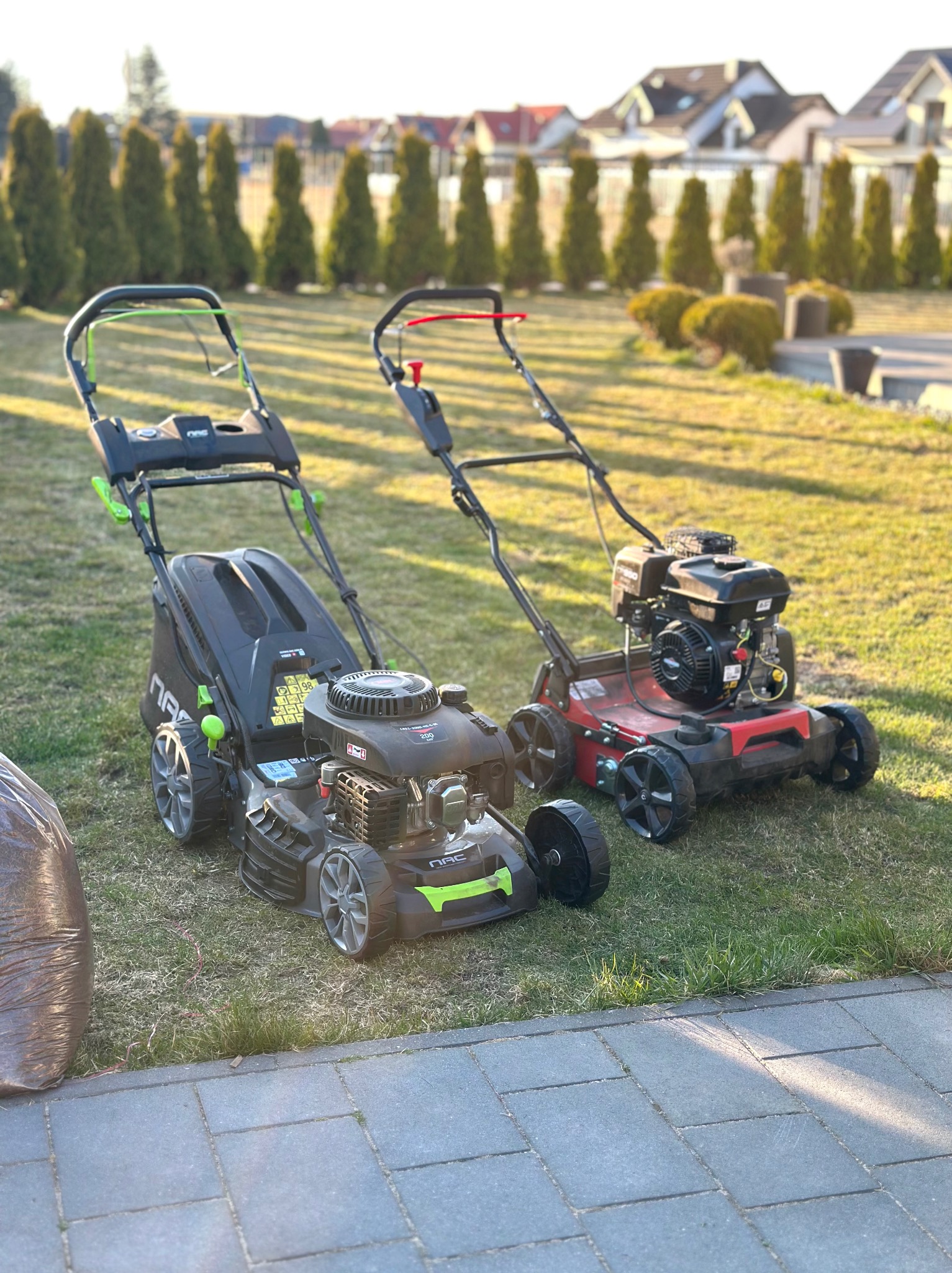 Dwie kosiarki do trawy (czarna i czerwona) stoją na trawniku przed domem z tujami w tle. Obok leży brązowy worek. Słoneczny dzień.