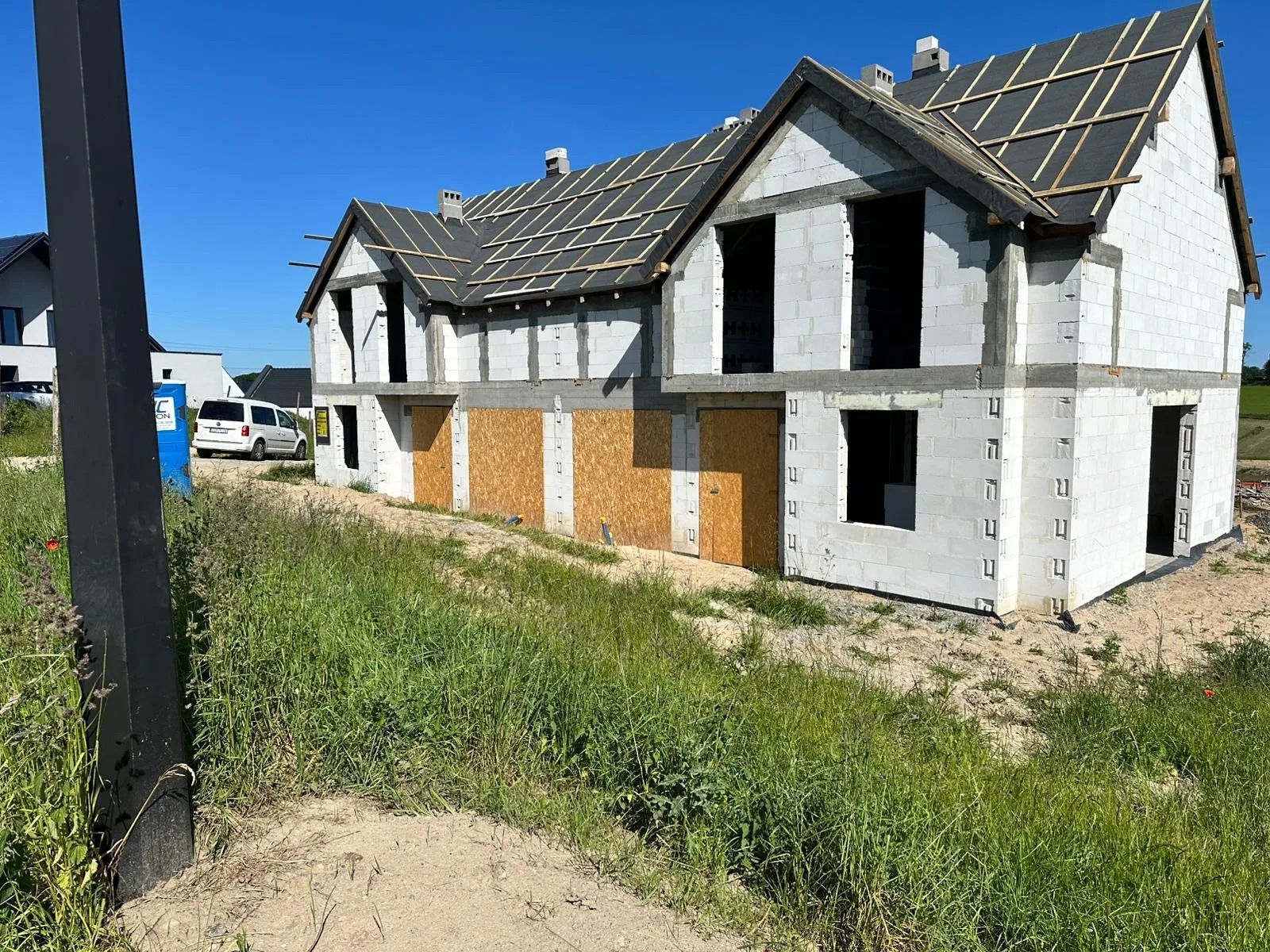 Surowy stan bliźniaka z białego bloczka, z deskami zamiast drzwi i dachem w budowie, na tle zieleni i błękitnego nieba. Widoczny samochód dostawczy.