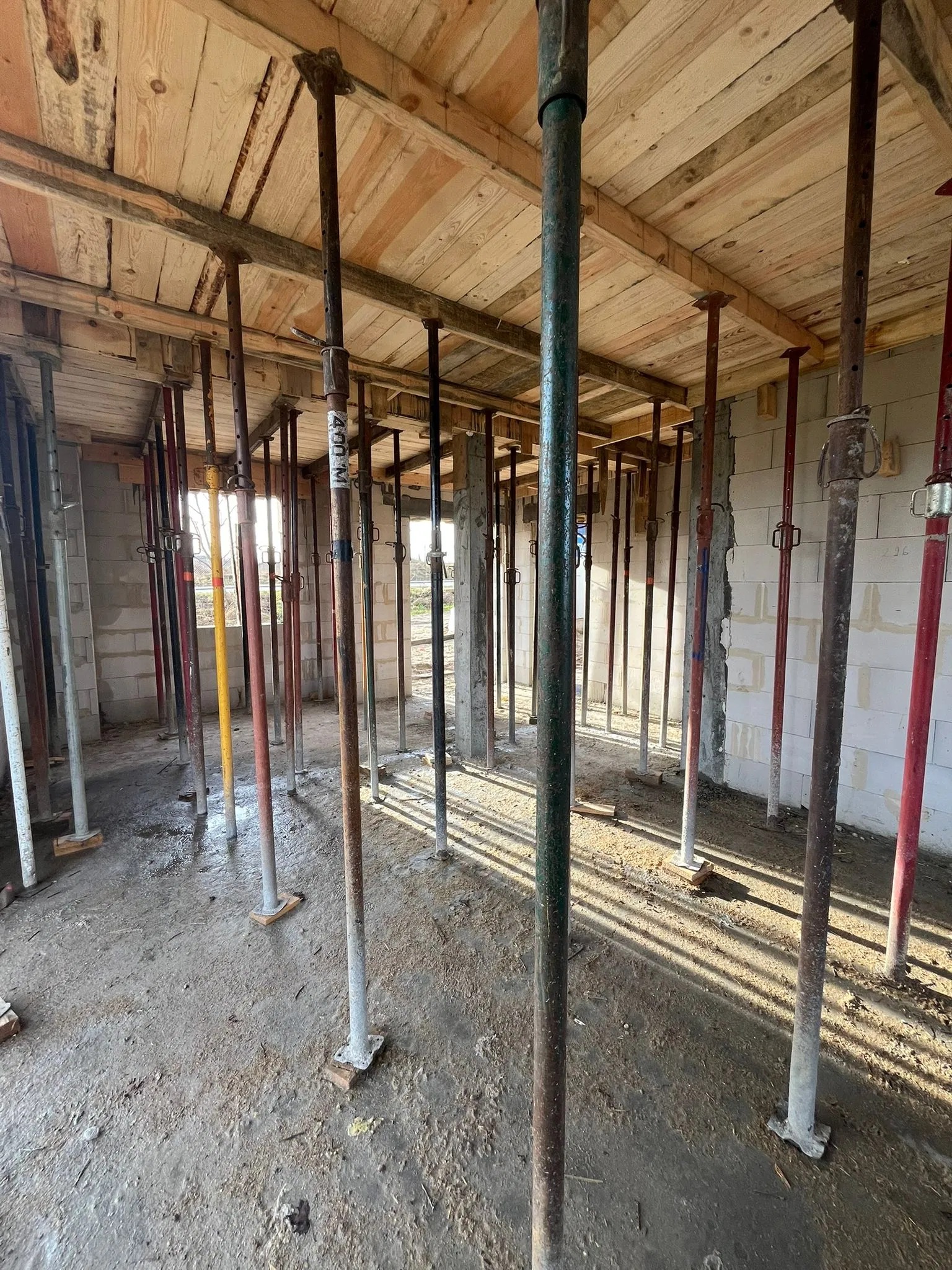 Wnętrze w trakcie budowy: drewniany strop podparty metalowymi stemplami, surowe ściany z bloczków, widoczne ślady betonu na podłodze, wpadające światło słoneczne.