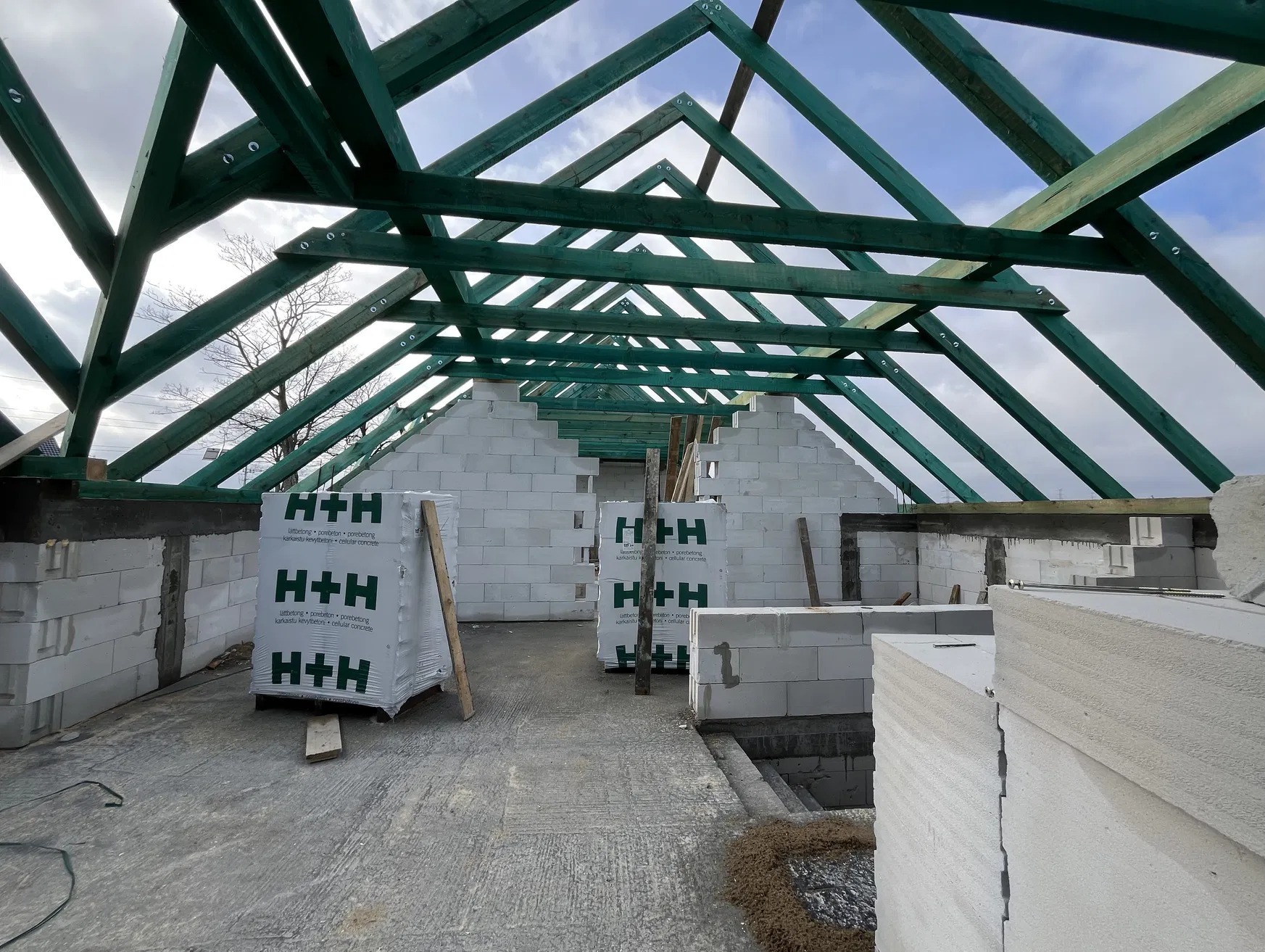 Wnętrze w trakcie budowy: ściany z betonu komórkowego H+H, zielona konstrukcja dachu z drewna. Widoczne materiały budowlane i surowy stan wykończenia.