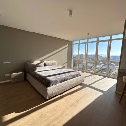 Oleksandr Matrofailo - Jasna sypialnia z dużym oknem i drewnianą podłogą. Beżowe ściany, łóżko z szarą narzutą. W odbiciu lustra widoczna osoba robiąca zdjęcie.