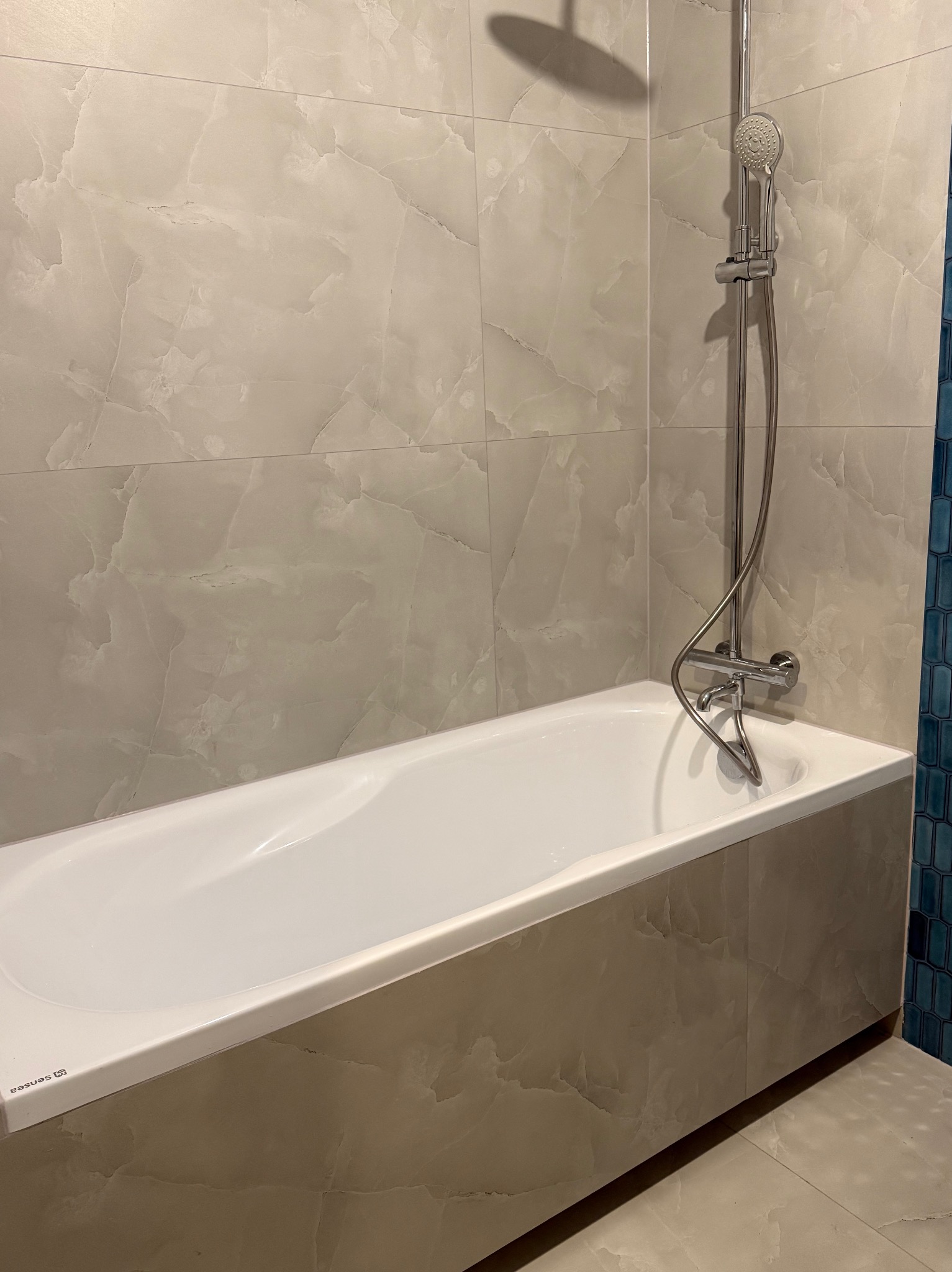 Biała wanna z obudową z płytek imitujących marmur, chromowana bateria i słuchawka prysznicowa na tle ściany wyłożonej płytkami w odcieniach beżu i błękitu.
