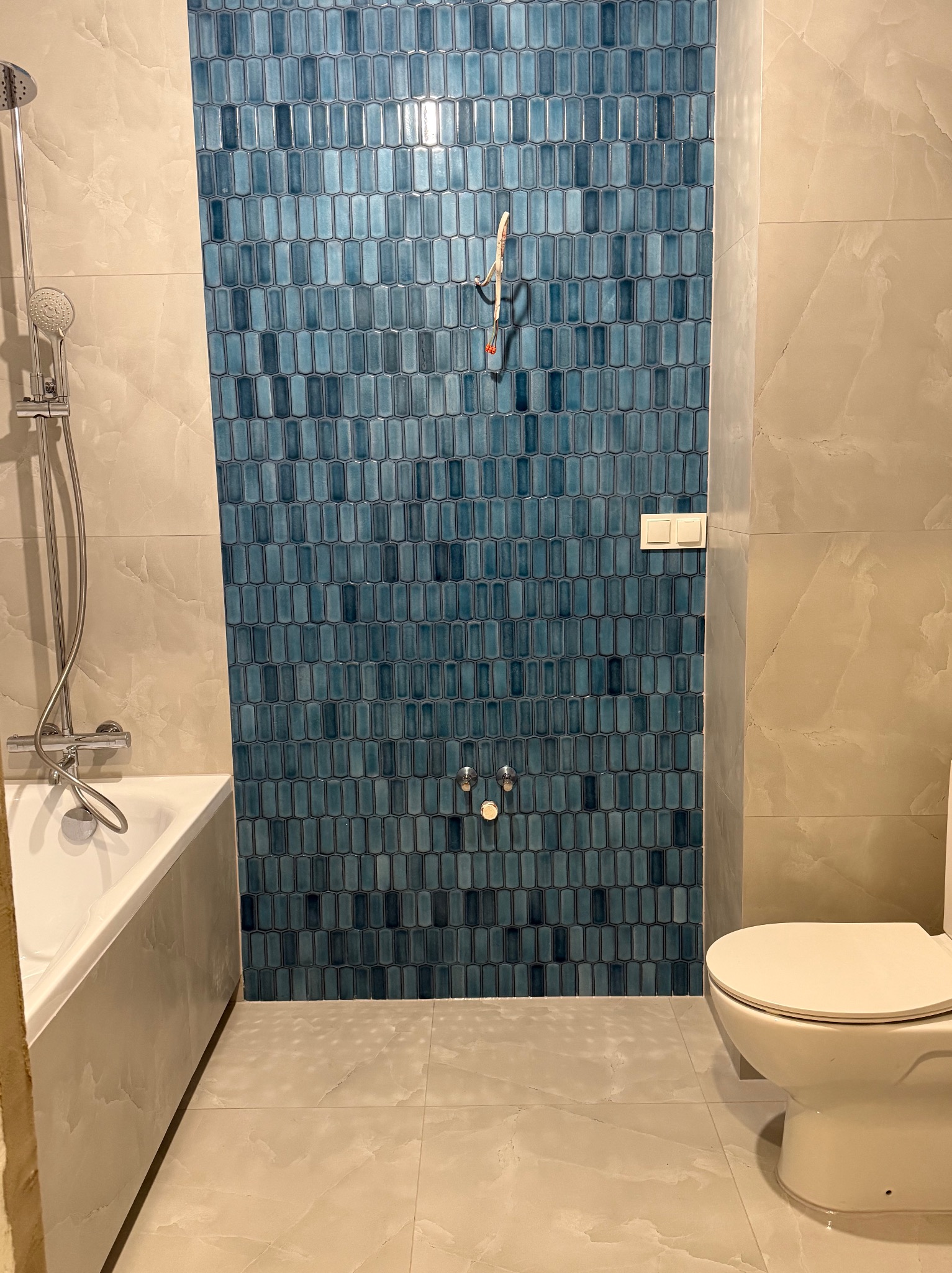 Wykończona łazienka z niebieską mozaiką na ścianie, beżowe płytki podłogowe i ścienne, biała wanna i toaleta. Widoczne elementy instalacji wodnej.