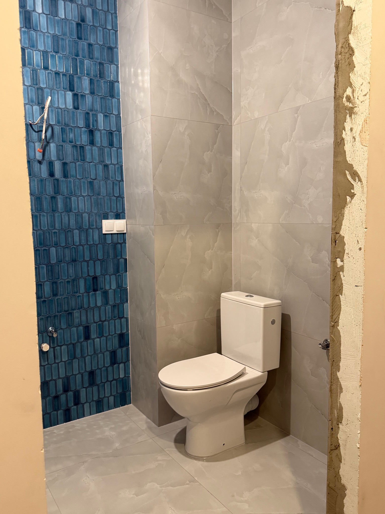 Wykończona łazienka z toaletą, ściany w szaro-białe marmurowe płytki i niebieską mozaikę. Widoczne niedokończone obramowanie drzwi.