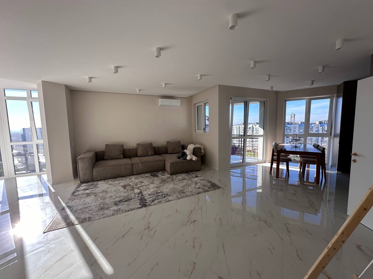 Jasne, przestronne wnętrze salonu w Gdyni z dużym narożnikiem, marmurową podłogą i widokiem na miasto przez okna balkonowe. Minimalistyczne oświetlenie sufitowe.