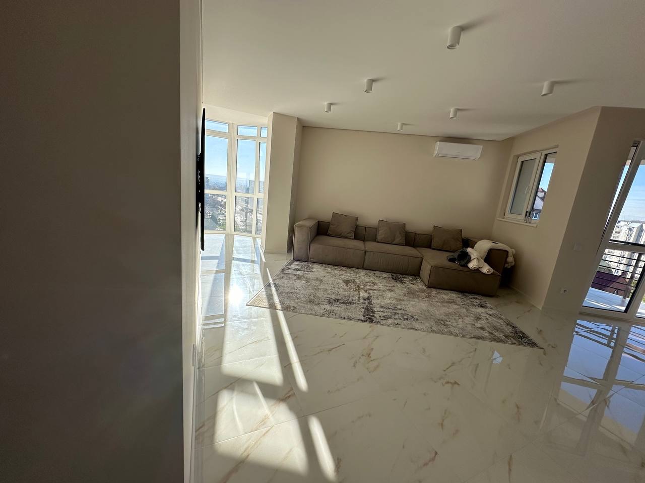 Jasne, przestronne wnętrze salonu z marmurową podłogą, narożną sofą i dużymi oknami z widokiem na miasto. Minimalistyczny design z klimatyzacją i nowoczesnym oświetleniem.