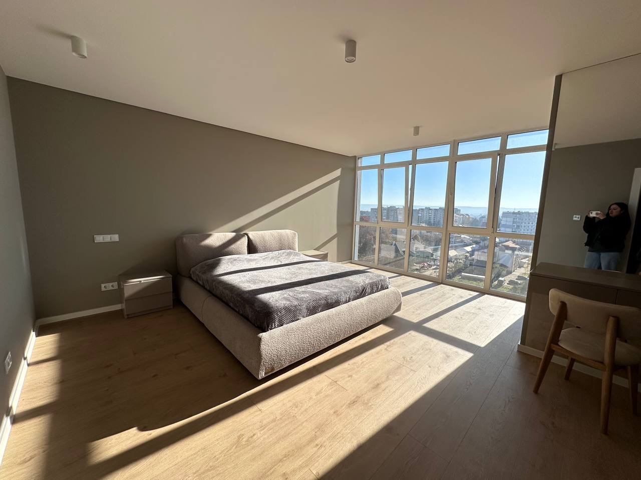 Jasna sypialnia z dużym oknem i drewnianą podłogą. Beżowe ściany, łóżko z szarą narzutą. W odbiciu lustra widoczna osoba robiąca zdjęcie.