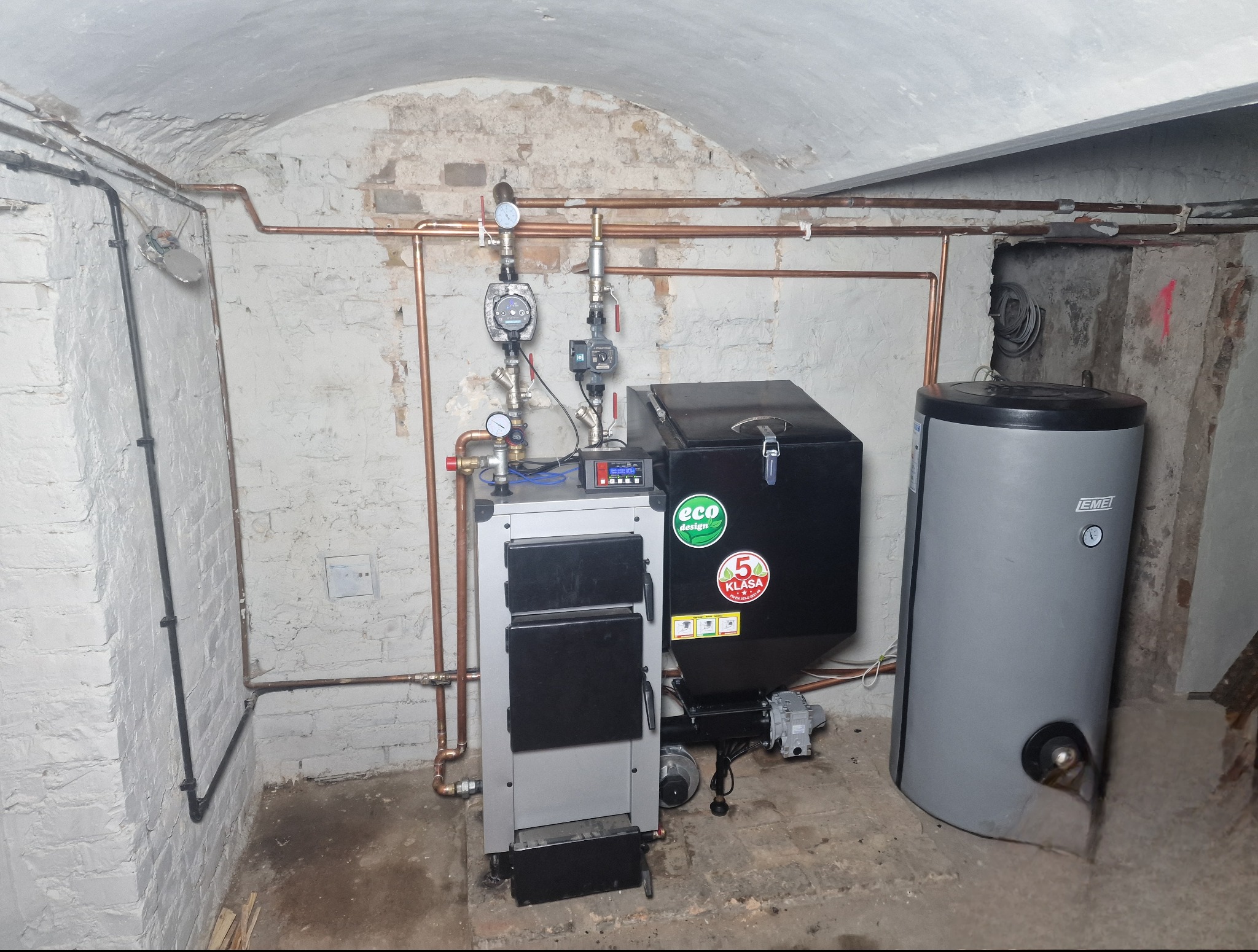 Nowoczesny piec CO z podajnikiem i zasobnikiem na paliwo, zainstalowany w piwnicy z ceglanym sklepieniem. Widoczne rury miedziane i osprzęt instalacyjny.