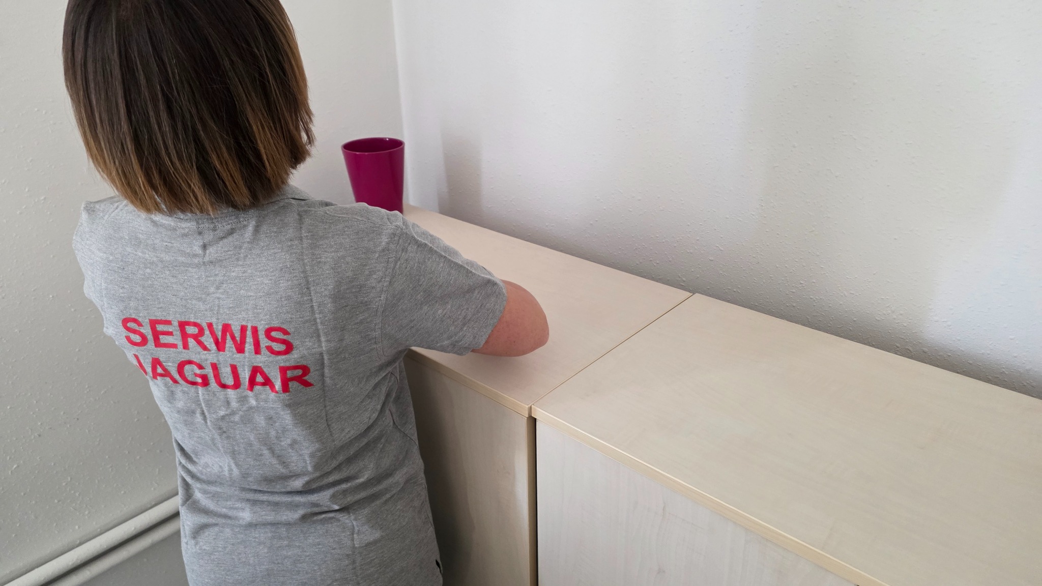 Osoba w szarej koszulce z napisem 'SERWIS JAGUAR' czyści jasny, drewniany mebel. Widoczny fioletowy kubek i fragment ściany z delikatną fakturą.
