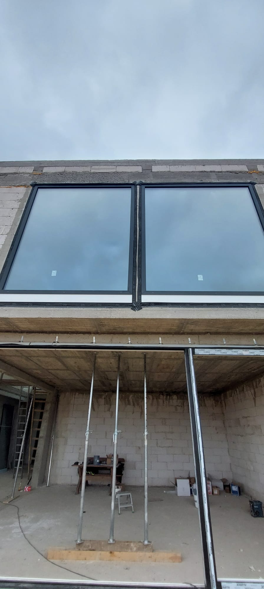 Dwa duże okna PCV z czarnymi ramami zamontowane w surowym stanie budynku. Widoczne podpory stropu i elementy konstrukcyjne. Ujęcie z dołu, pochmurne niebo w tle.