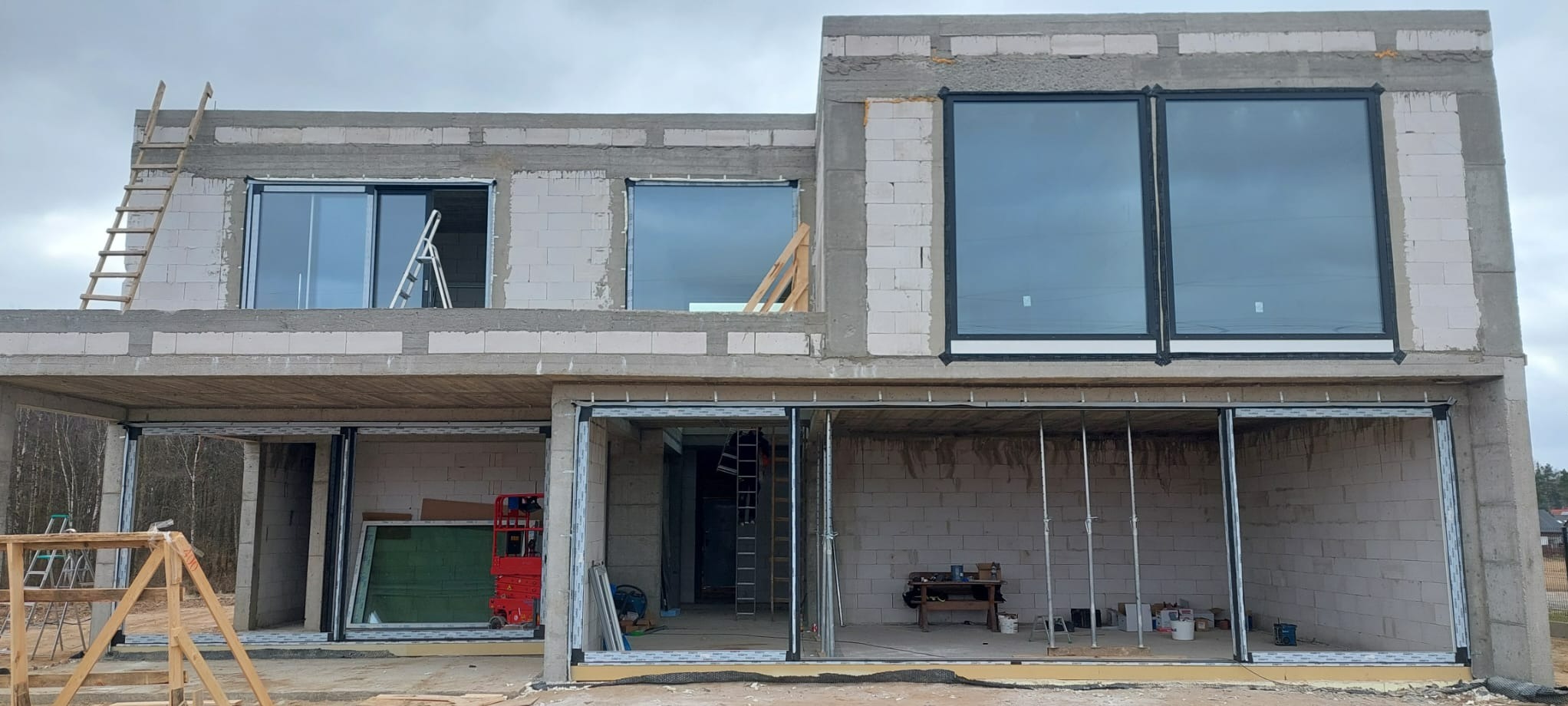 Montaż okien i drzwi przesuwnych w surowym, dwukondygnacyjnym domu z betonu komórkowego. Widoczne drabiny i narzędzia budowlane.