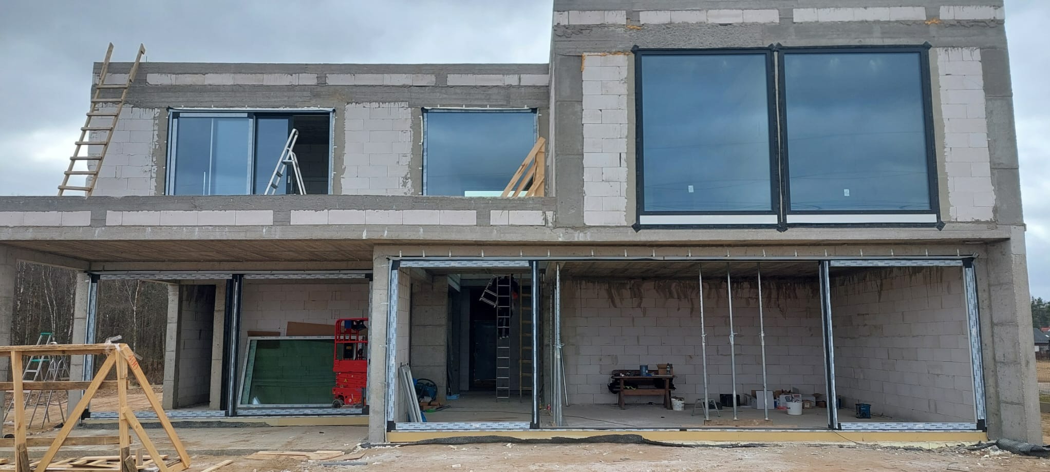 Surowy stan budynku w trakcie montażu okien i drzwi przesuwnych. Widoczne rusztowania i drabiny. Okna z czarnymi ramami odbijają pochmurne niebo.