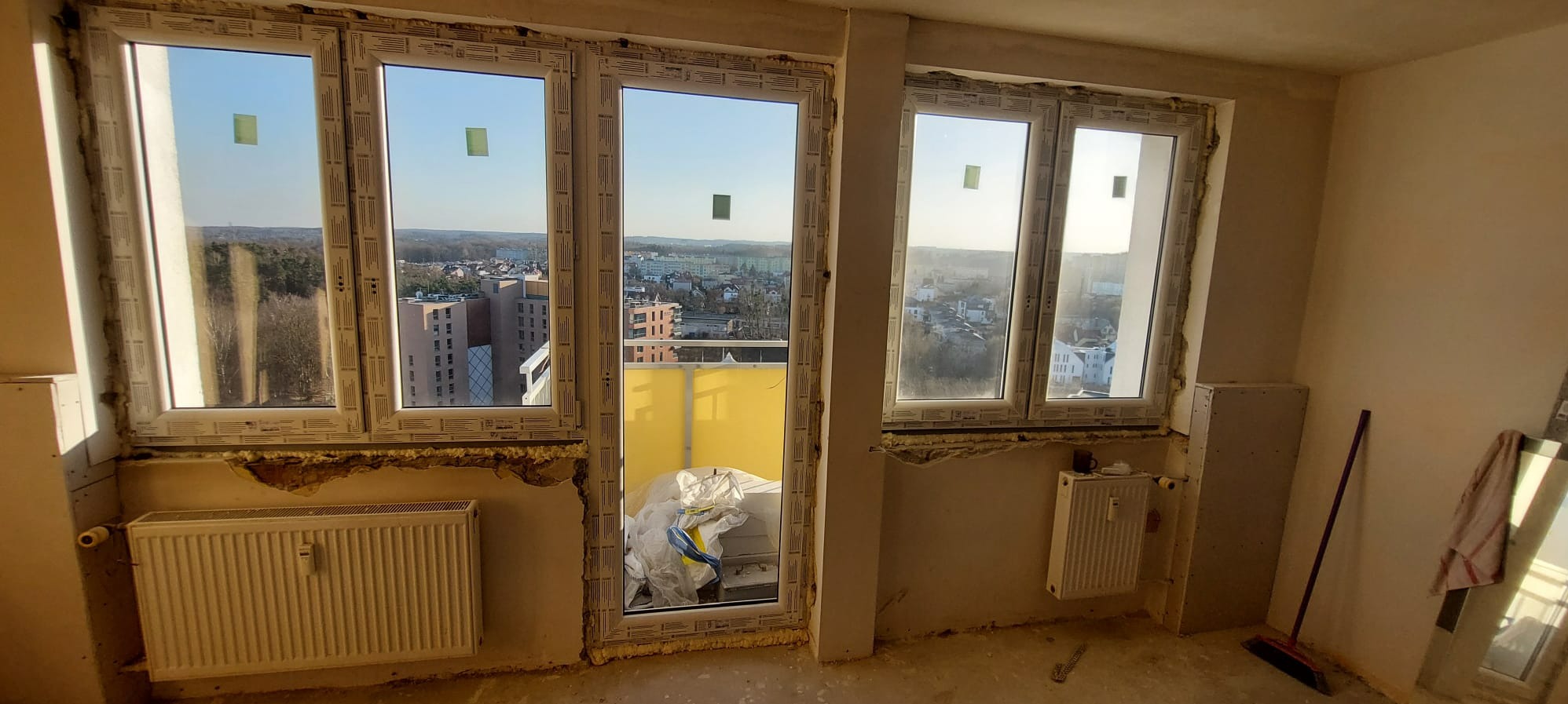 Montaż okien PCV w surowym wnętrzu z widokiem na miasto. Widoczne grzejniki i ślady prac wykończeniowych. Dwa okna i drzwi balkonowe z zielonymi naklejkami.