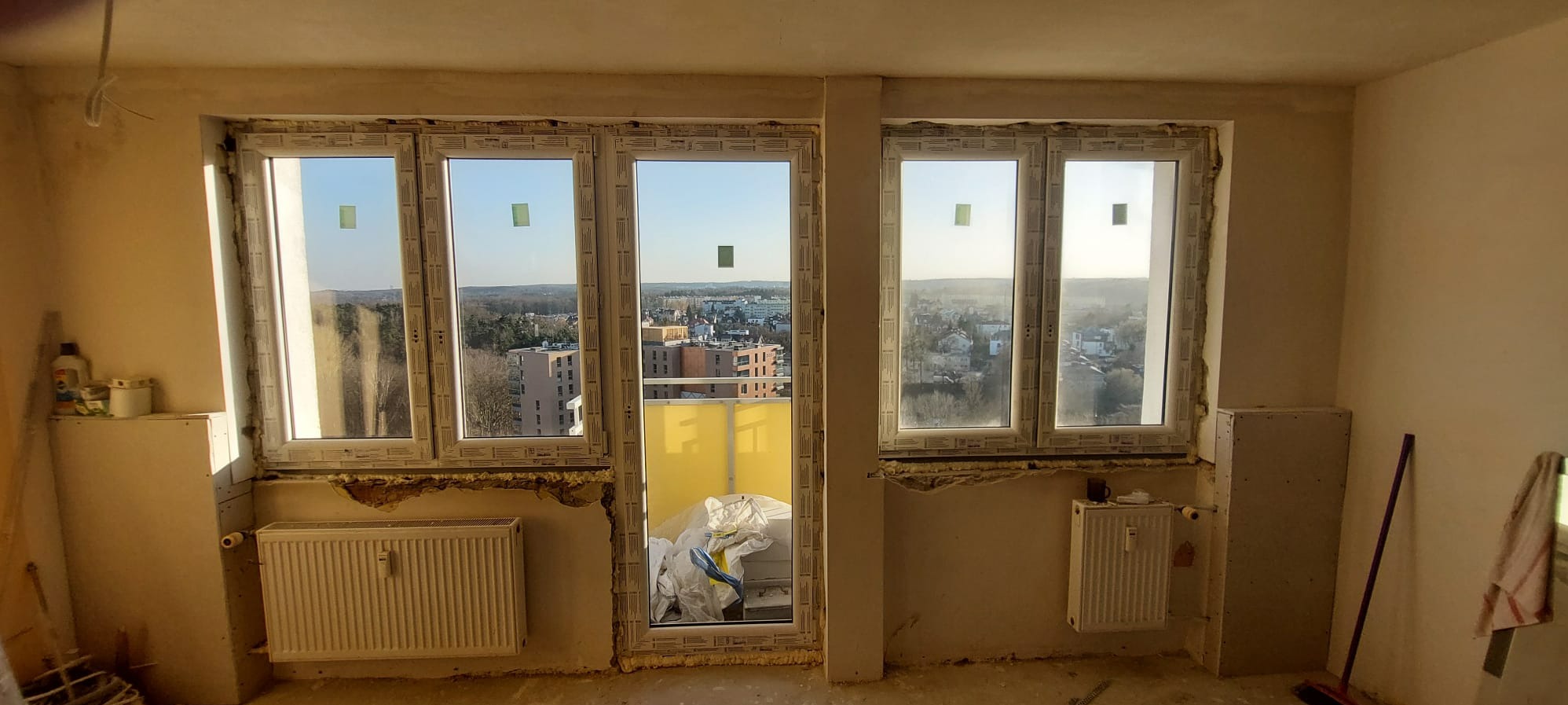 Wnętrze z dwoma oknami PCV i drzwiami balkonowymi, widok na miasto. Ślady montażu, grzejniki pod oknami, surowy stan wykończenia ścian. Dzień słoneczny.