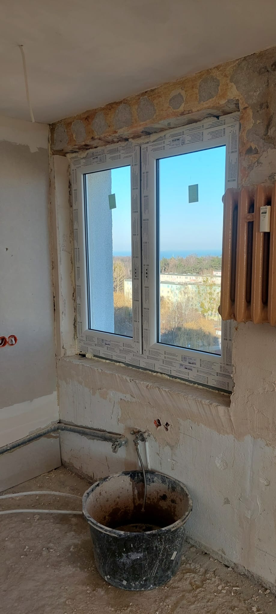 Nowe, białe okno PCV z widokiem na zewnątrz, zamontowane w surowym wnętrzu. Widoczne ślady po remoncie, grzejnik i wiadro. Okno z zielonymi naklejkami.