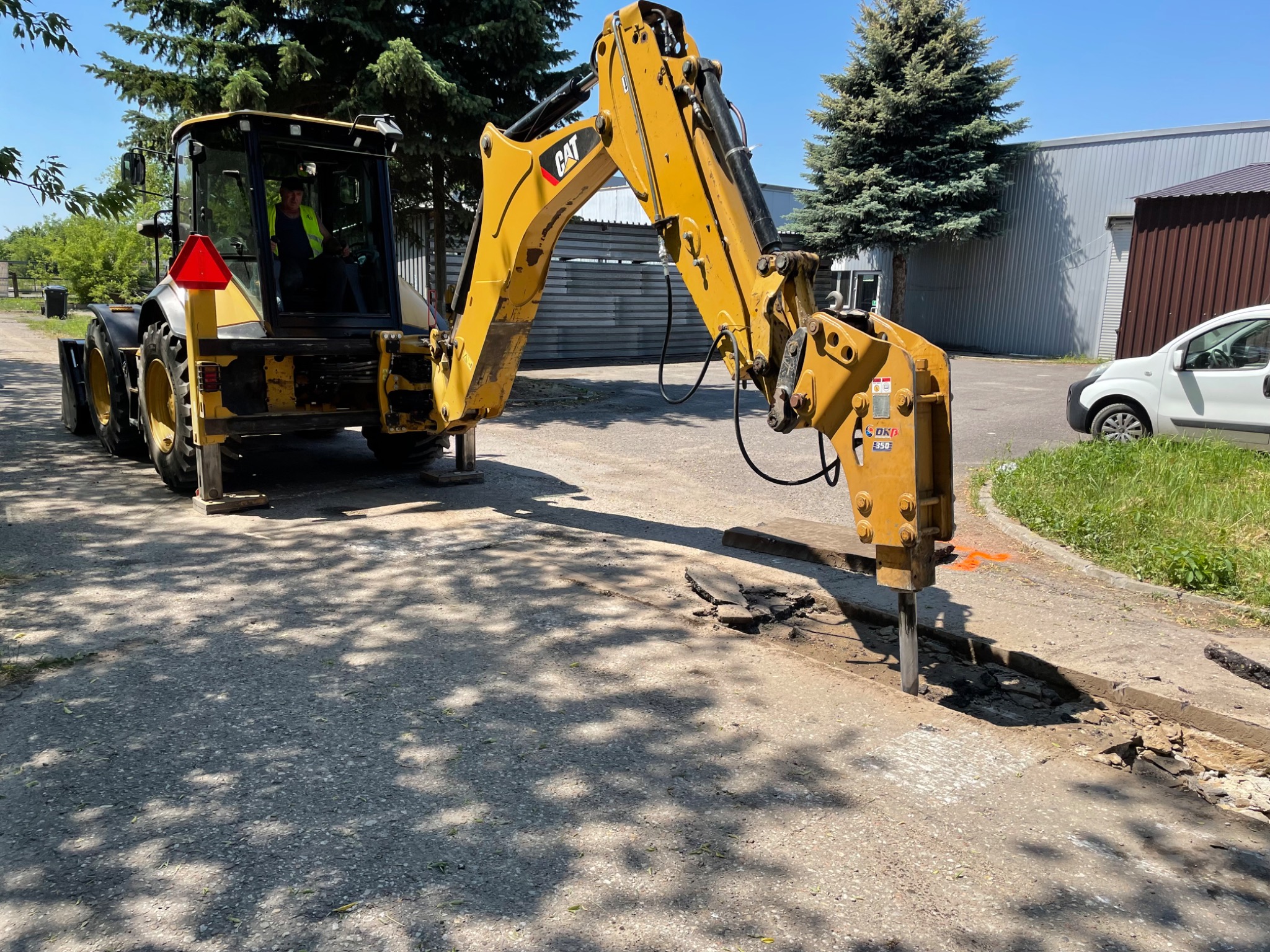 Żółta koparka CAT z pneumatycznym młotem rozbija betonowy chodnik na zewnątrz, w tle budynek i drzewa. Operator w kabinie. Drobne uszkodzenia chodnika.
