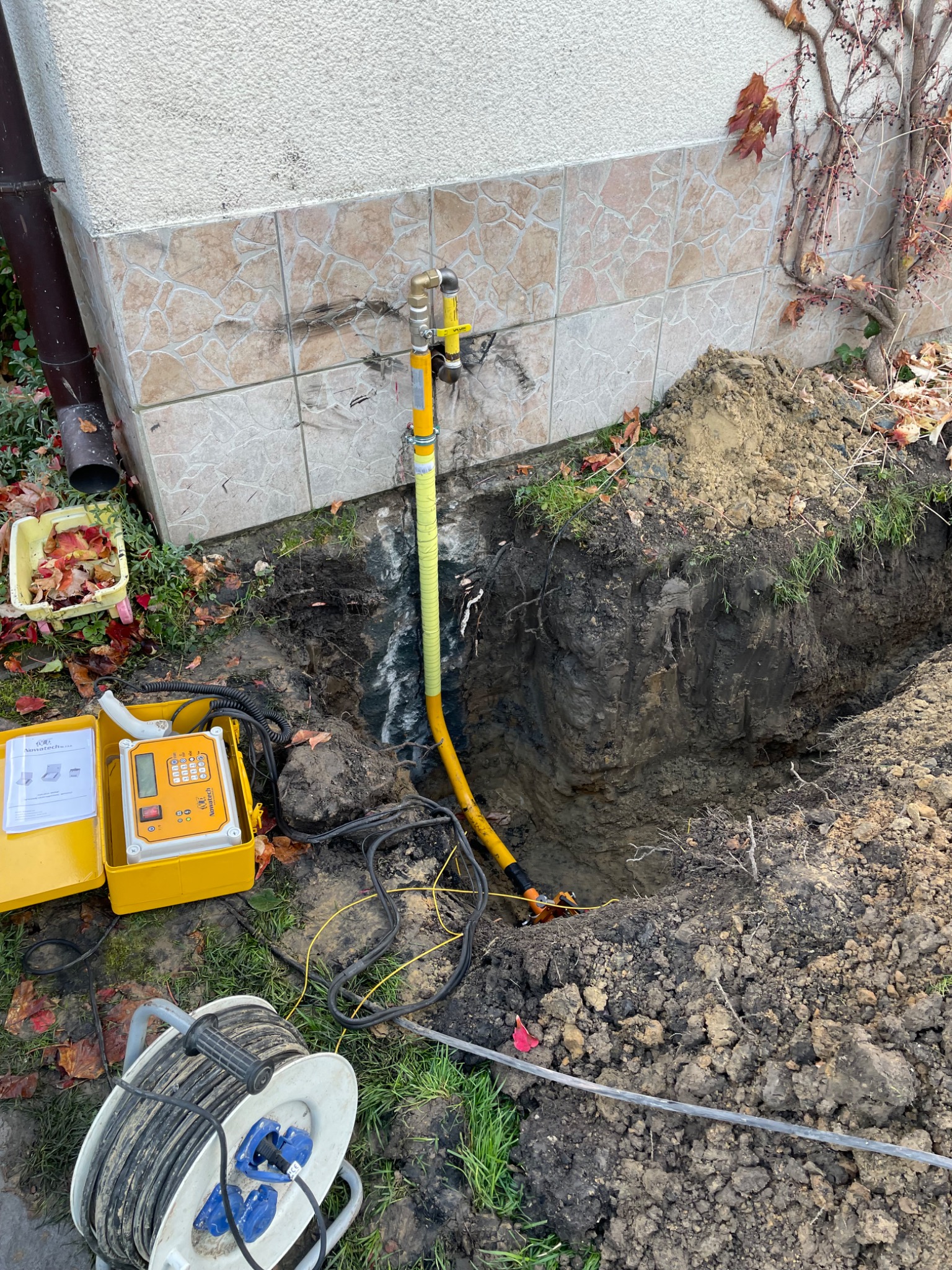 Żółta rura gazowa w wykopie przy ścianie budynku, obok sprzęt pomiarowy. Prace ziemne i instalacyjne w toku, widoczne narzędzia i elementy instalacji.