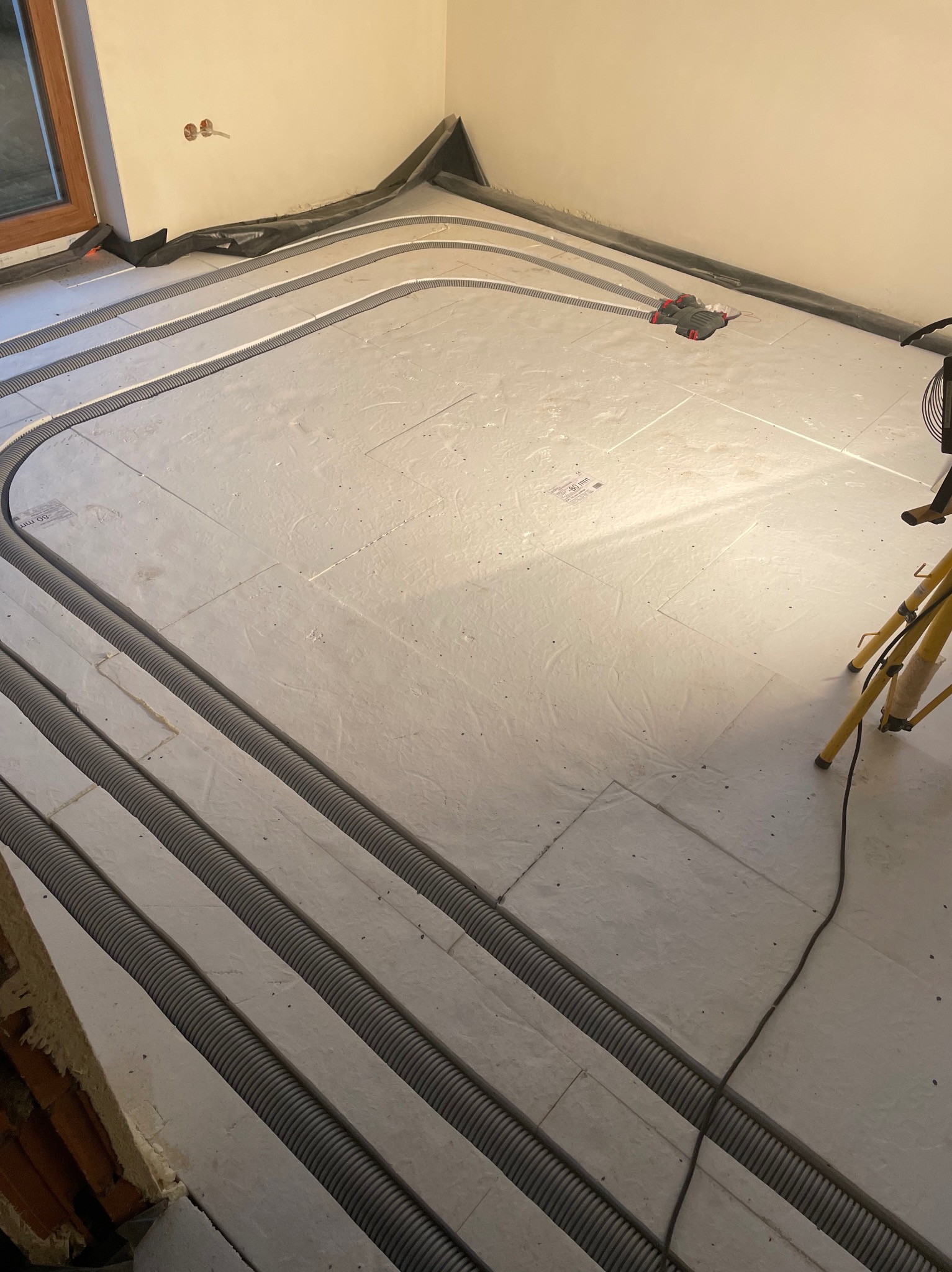 Ułożone rury ogrzewania podłogowego na izolacji, widoczne narzędzia i fragment ściany. Etap prac przed wylaniem posadzki. Szare rury ułożone w pętle.