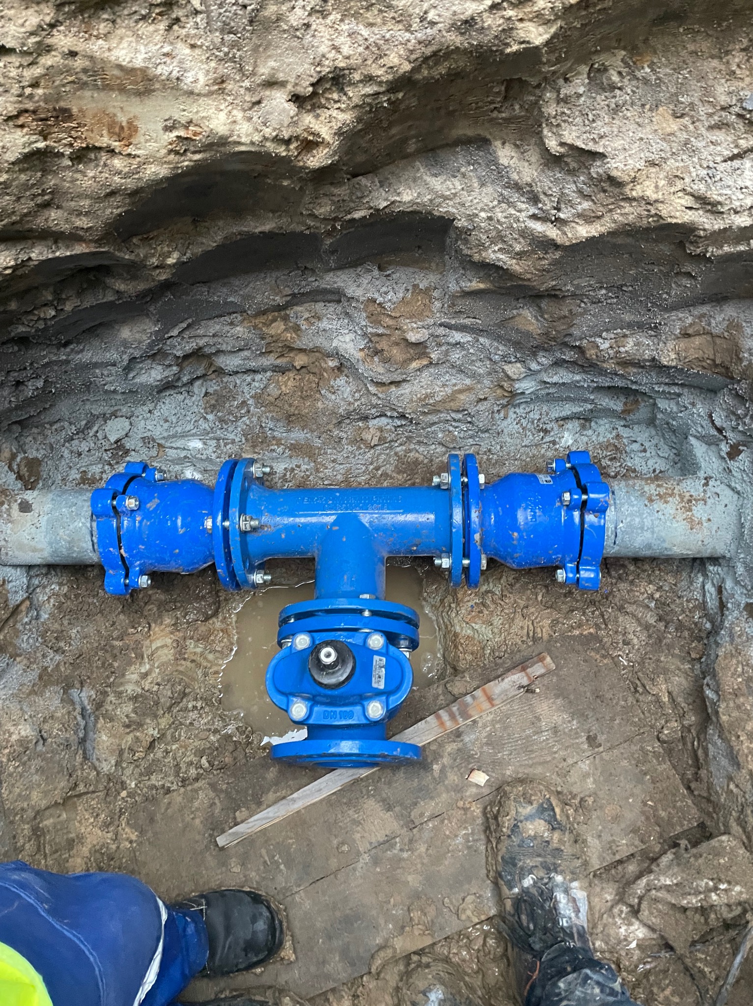 Niebieski zawór z przyłączami zamontowany w wykopie ziemnym. Widoczne fragmenty rur, ziemia i buty osoby wykonującej prace. Instalacja wodociągowa.