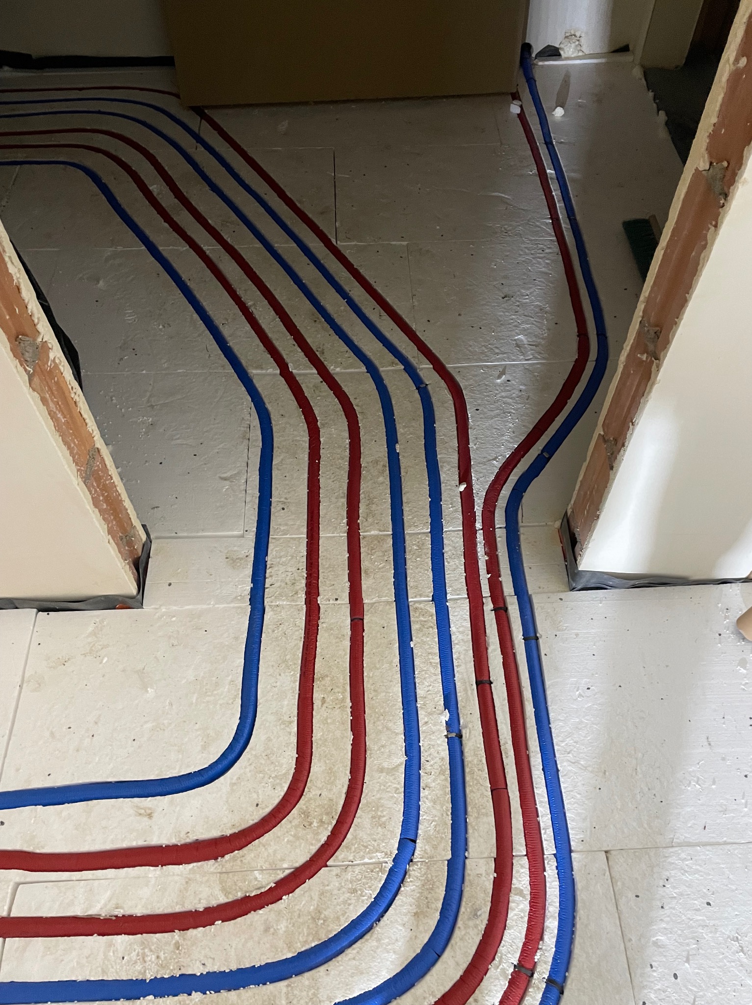 Ułożone, czerwono-niebieskie rury instalacji podłogowej na jasnej posadzce, widok z góry. Rury prowadzone równolegle, zakręcają w lewo i prawo.