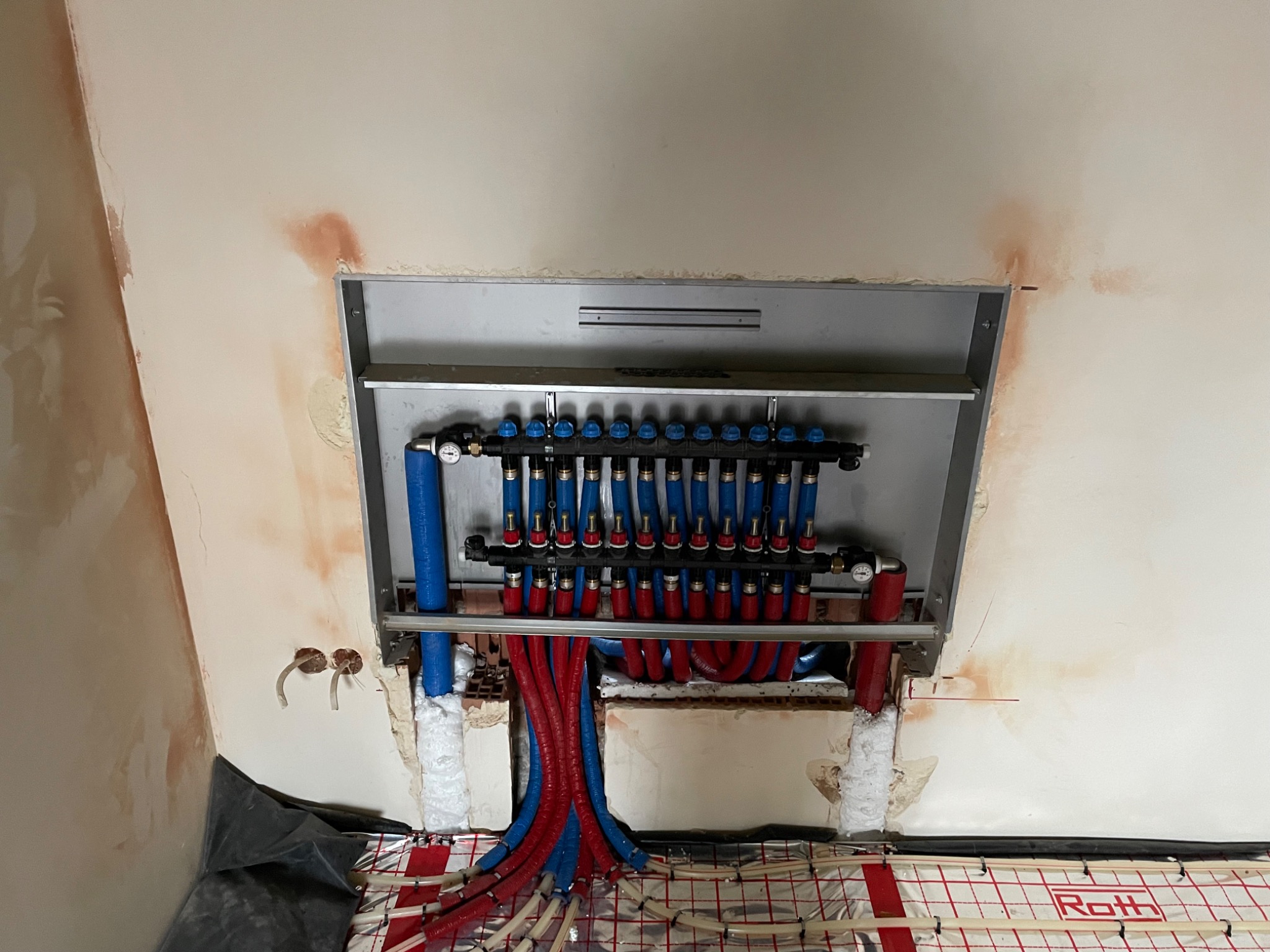 Rozdzielacz instalacji ogrzewania podłogowego Roth, z niebieskimi i czerwonymi rurami, zamontowany w ścianie na tle folii izolacyjnej i surowego tynku.