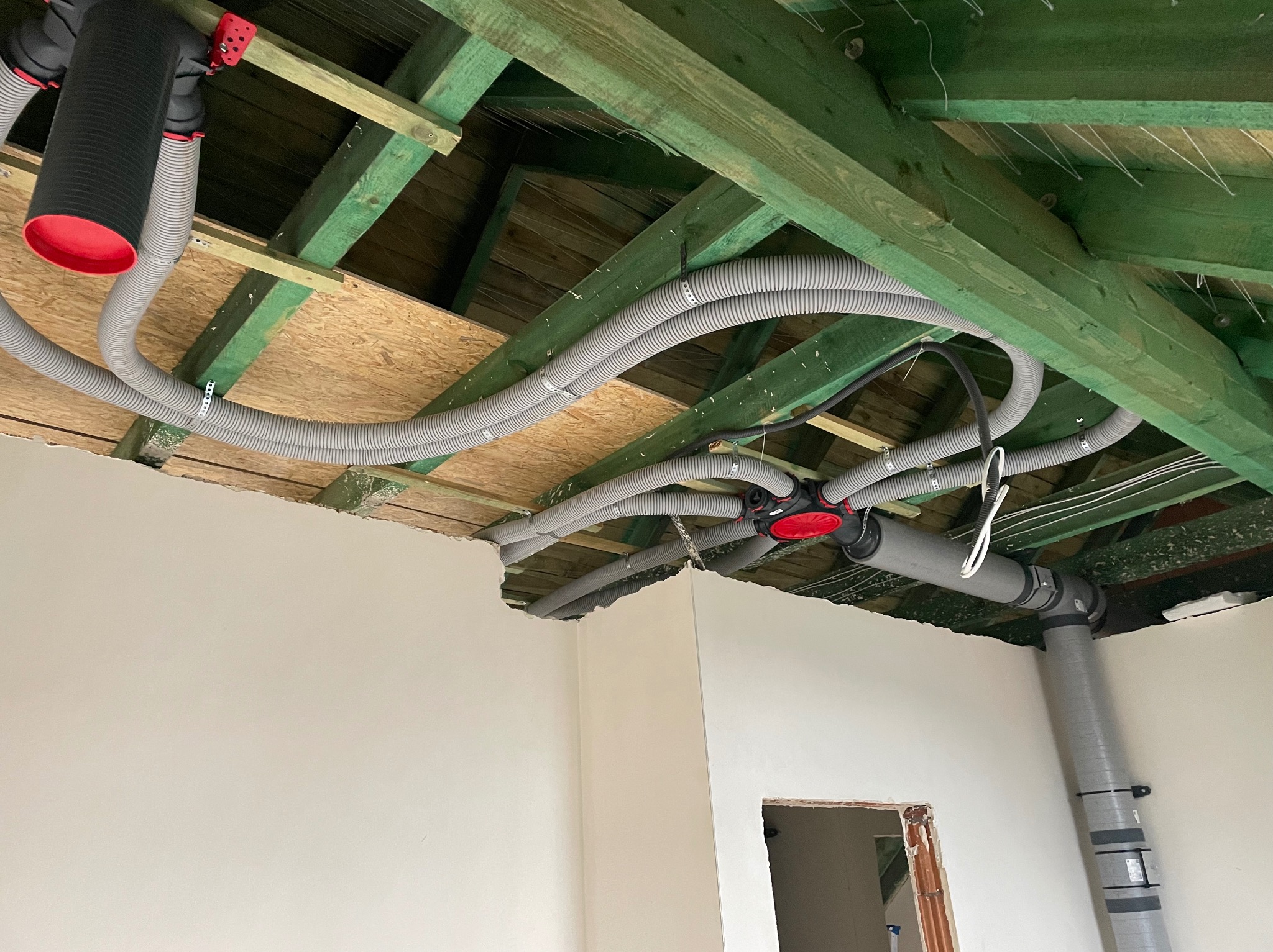 Szare, elastyczne rury wentylacyjne z czerwonymi elementami łączącymi, zamontowane pod zielonymi belkami stropu w surowym wnętrzu. Widoczny fragment ściany i otwór drzwiowy.