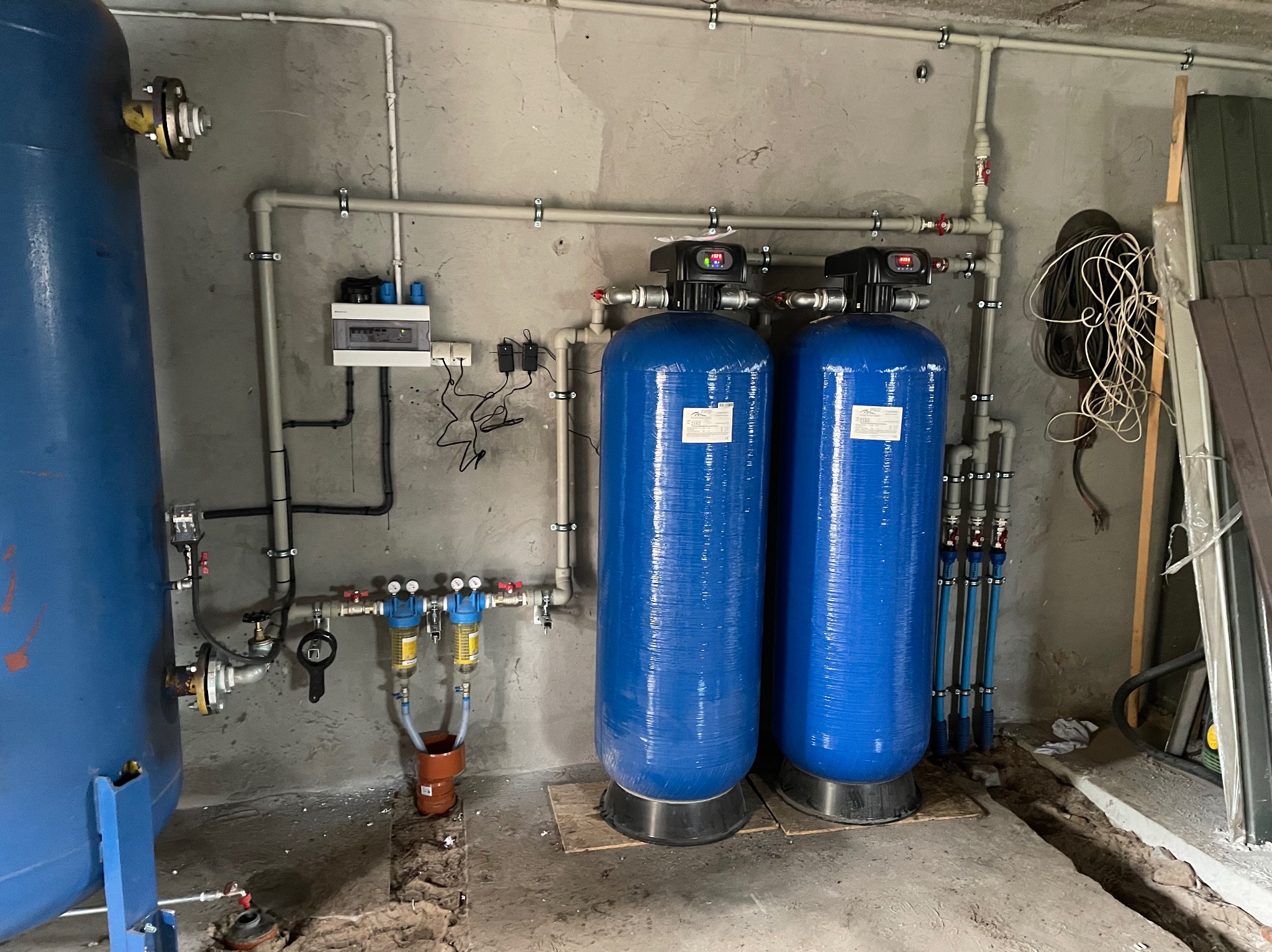 Dwa niebieskie filtry wody z manometrami i rurami na tle surowej ściany. Widoczna instalacja wodna z zaworami, rurami i elementami sterującymi.