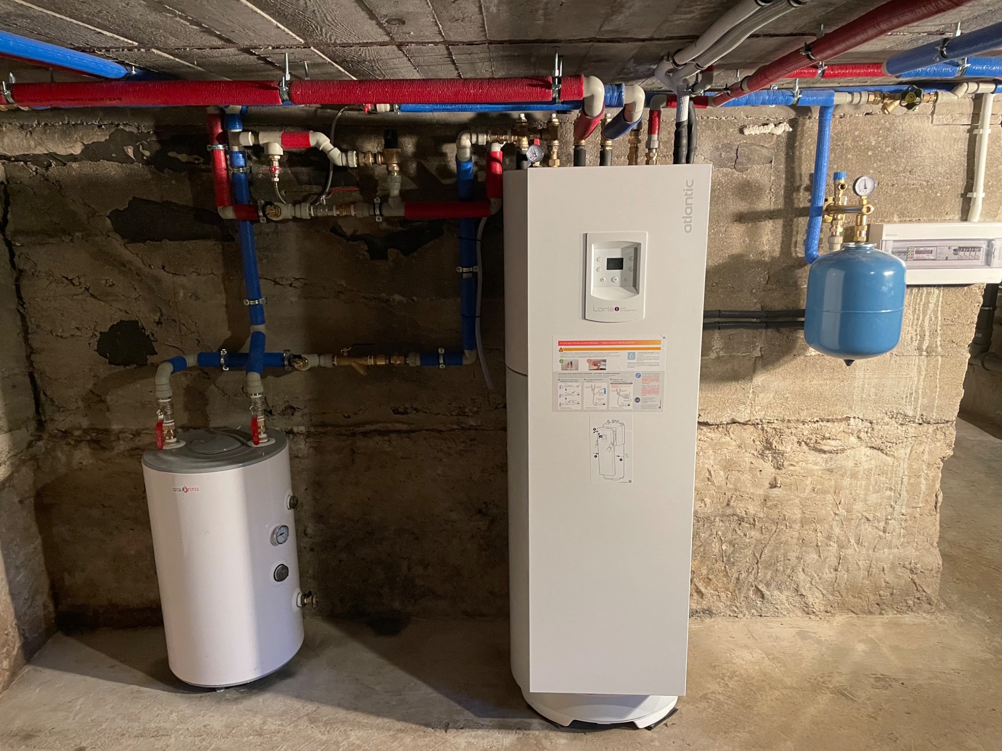 Biały, nowoczesny piec Atlantic z instalacją CO w piwnicy o surowych ścianach. Widoczne rury w izolacji, zbiornik wyrównawczy i rozdzielnica elektryczna.