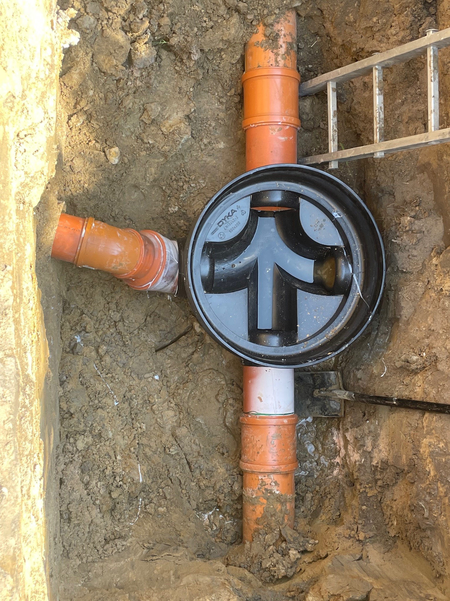 Studzienka kanalizacyjna z rurami w wykopie, widok z góry. Pomarańczowe rury połączone z czarną studzienką. Częściowo widoczna drabina.