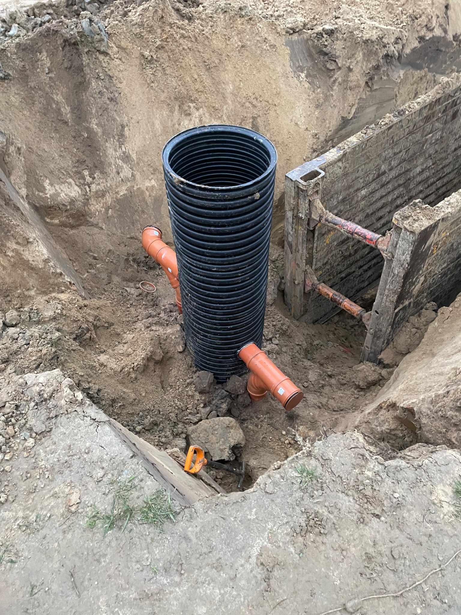 Wykop z zabezpieczeniami, w nim pionowa rura karbowana i dwie pomarańczowe rury kanalizacyjne. Widoczne złącza i fragmenty ziemi. Prace ziemne w toku.